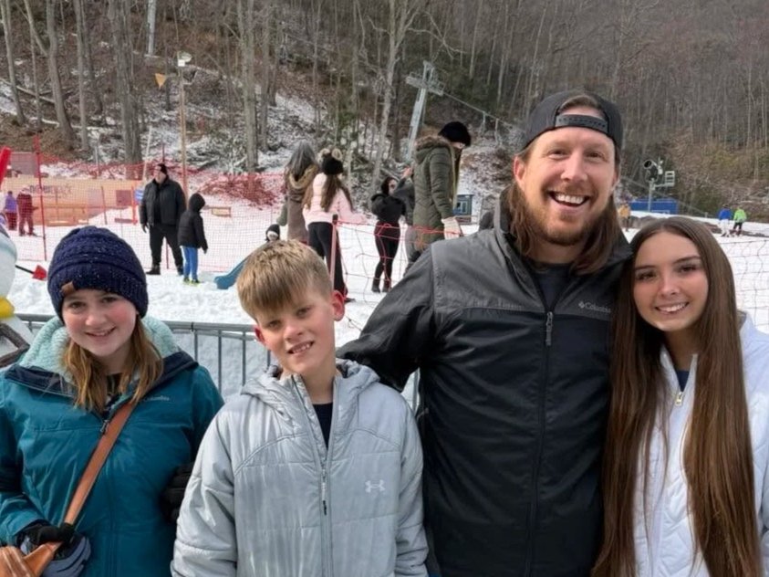 A group of four people, two girls and two boys, standing outdoors in a snowy area with ski slopes and other people in the background. They are smiling and dressed in winter jackets.