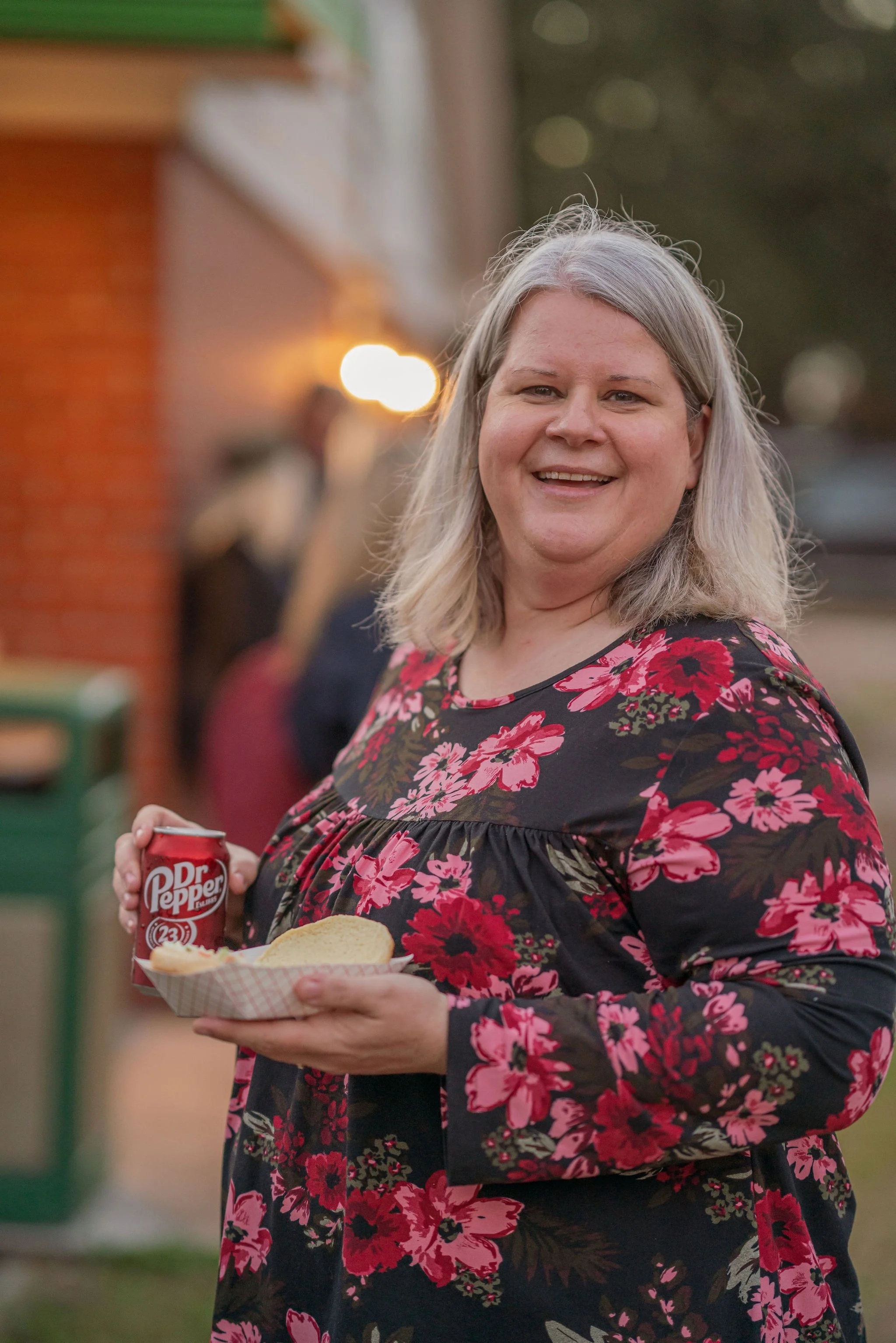A woman with gray hair, wearing a black dress with red and pink flowers, holding a tray with a hot dog and a can of Dr. Pepper at an outdoor gathering during sunset.