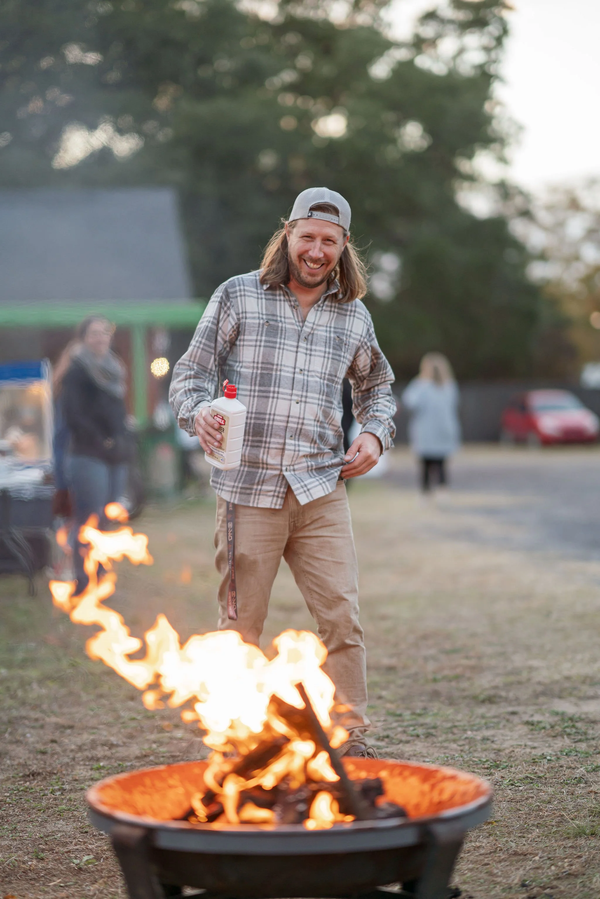 A smiling man with long hair, wearing a gray baseball cap and plaid shirt, standing outdoors at sunset near a fire pit, holding a bottle of mustard, with blurred background of other people and trees.