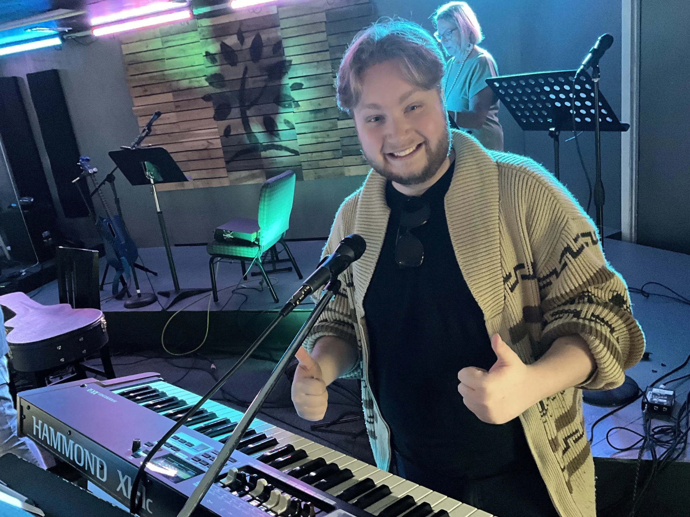 A young man in a yellow patterned cardigan giving two thumbs up while standing in front of a musical keyboard and microphone on stage. Behind him, a woman with glasses is looking down, with a wood-paneled wall and colorful stage lighting as backgroun