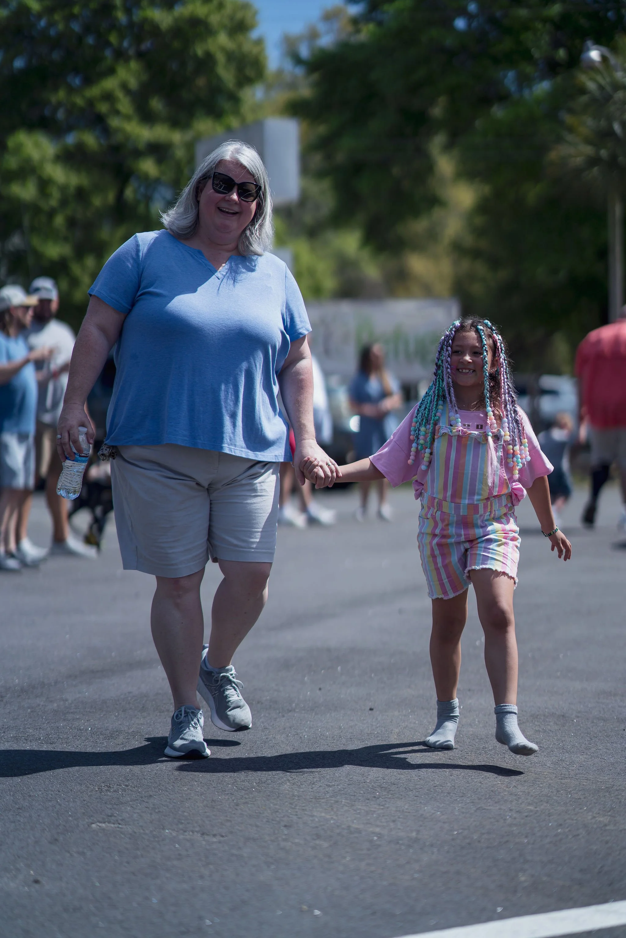 An older woman with gray hair, sunglasses, and a blue t-shirt holding hands with a young girl with colorful braids, wearing a striped outfit and boots, walking outdoors during a sunny day in a public area with trees and other people in the background