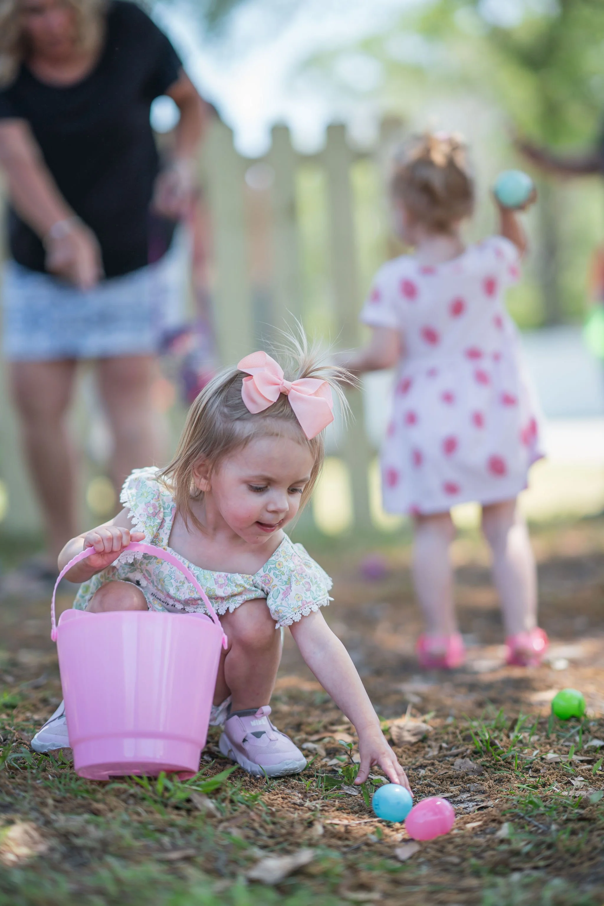 A young girl with a pink bow in her hair, wearing a floral dress, kneeling on the ground during an Easter egg hunt, reaching for plastic eggs with a pink bucket nearby. In the background, another girl in a white dress with pink spots and pink shoes i