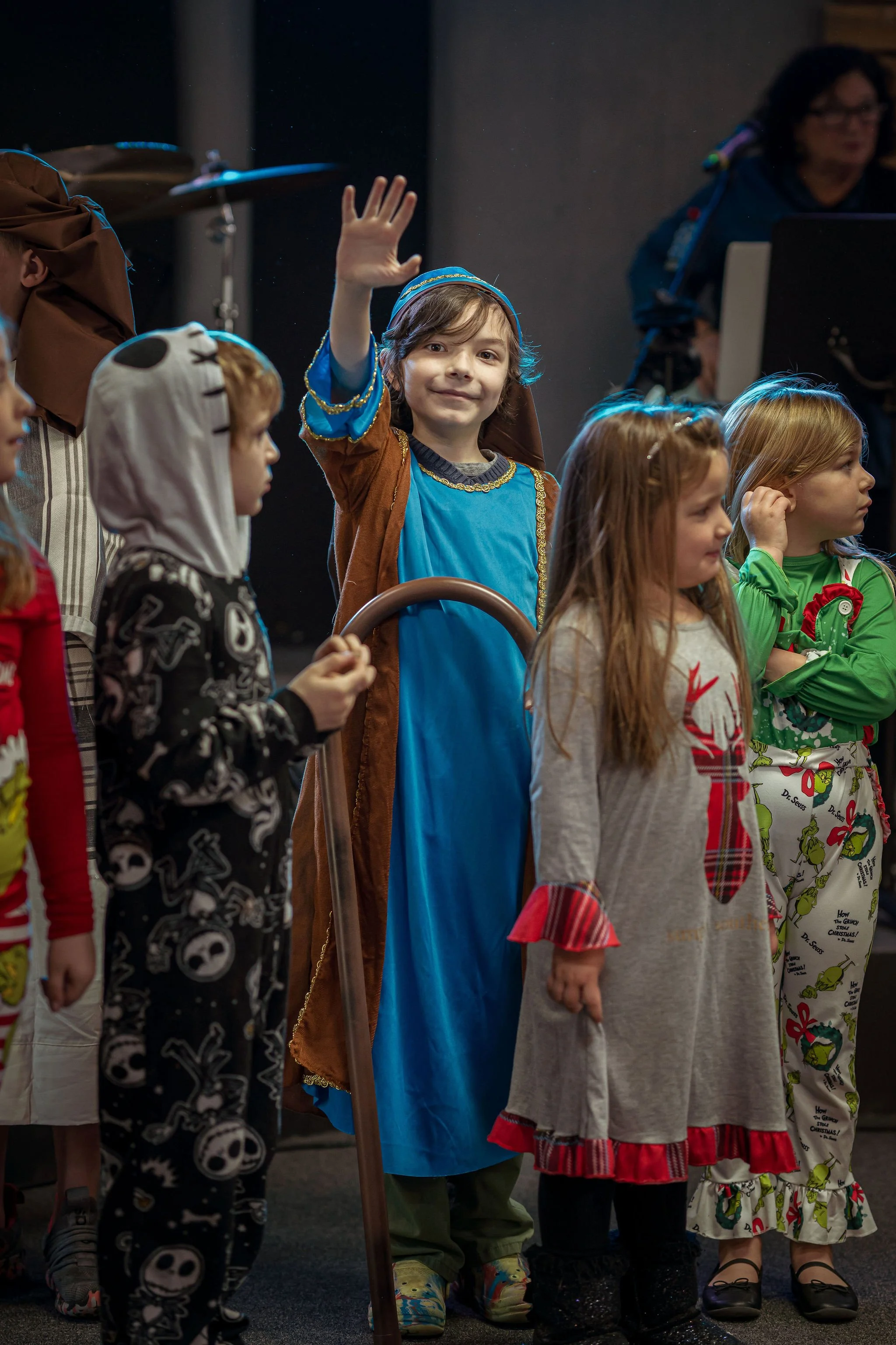 Children dressed in costumes, including a child in a blue and brown costume waving, standing in a line during a festive event.