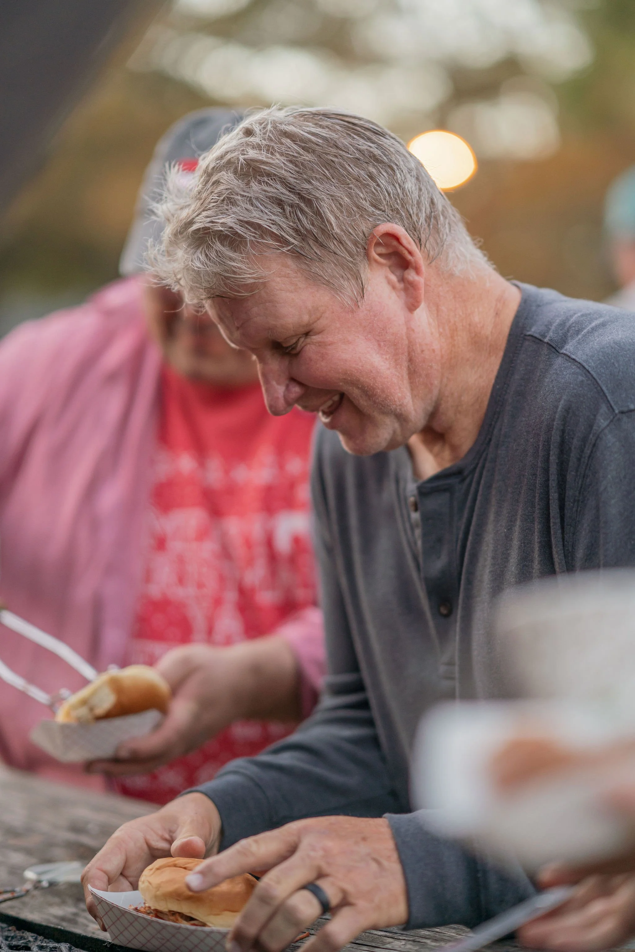 A man and a woman are preparing food at a table outdoors, with the man holding a hot dog bun and the woman serving food from a tray, both smiling and enjoying their time.