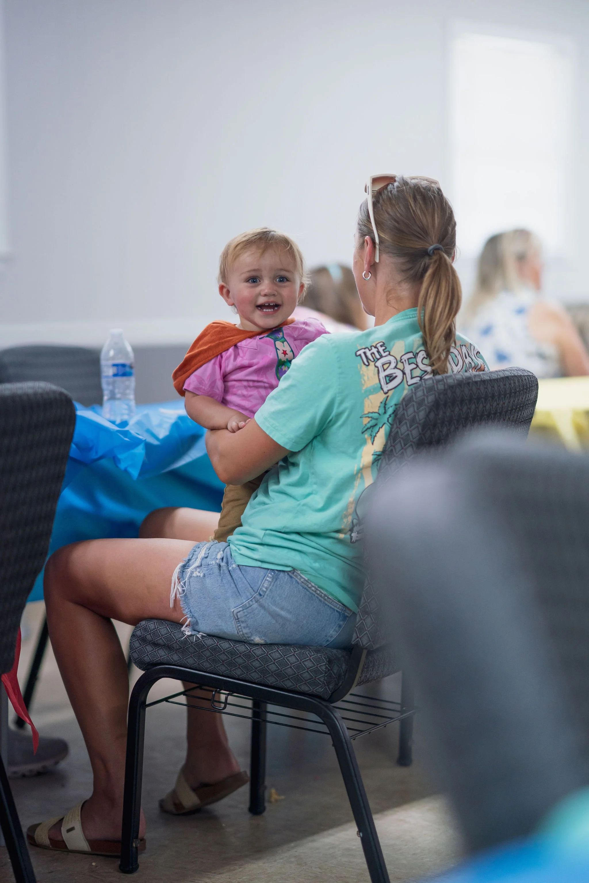 A woman with a ponytail sitting on a chair, holding a young boy with a concerned or upset expression. The woman is wearing sunglasses on her head, a turquoise shirt with a tie-dye pattern, and denim shorts. The boy is wearing a pink shirt with a colo