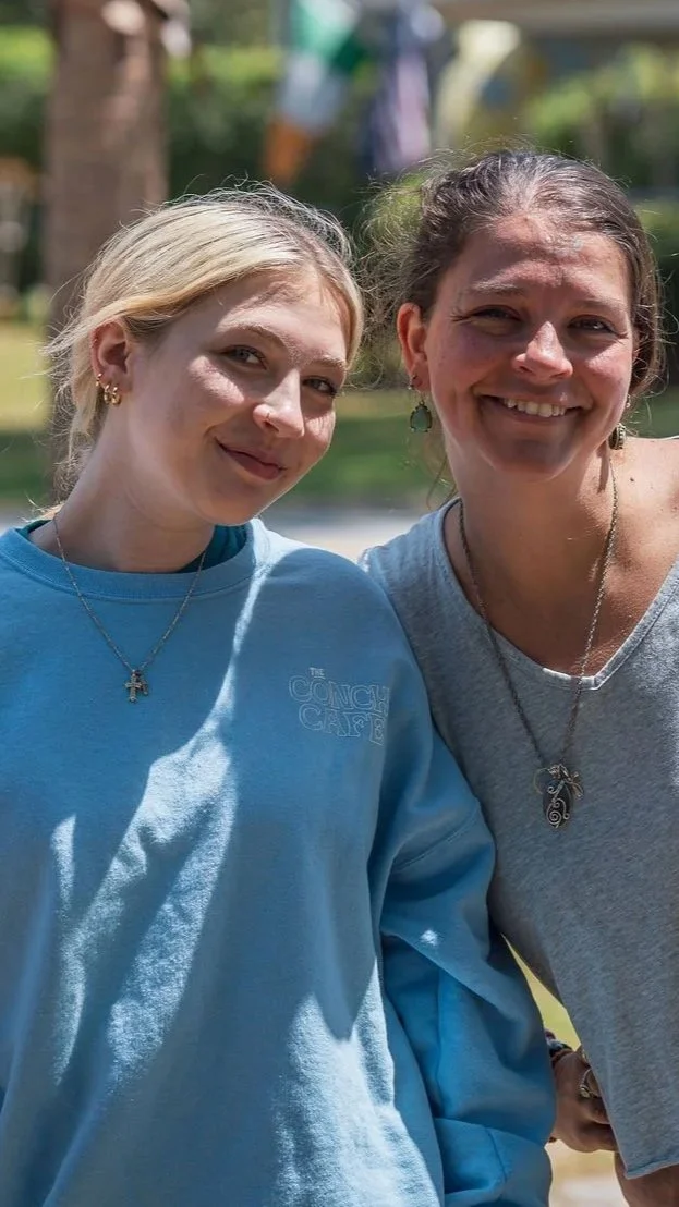 Two women smiling outdoors, one wearing a light blue sweatshirt and the other a gray T-shirt, with trees and a pond in the background.