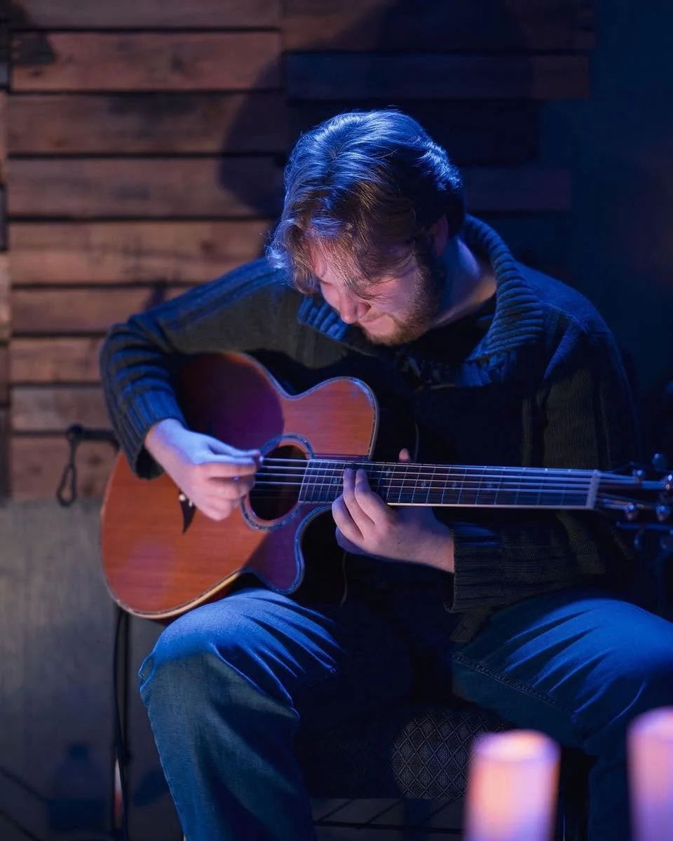 A young man with brown hair and a beard playing an acoustic guitar in a dimly lit room with wooden walls.