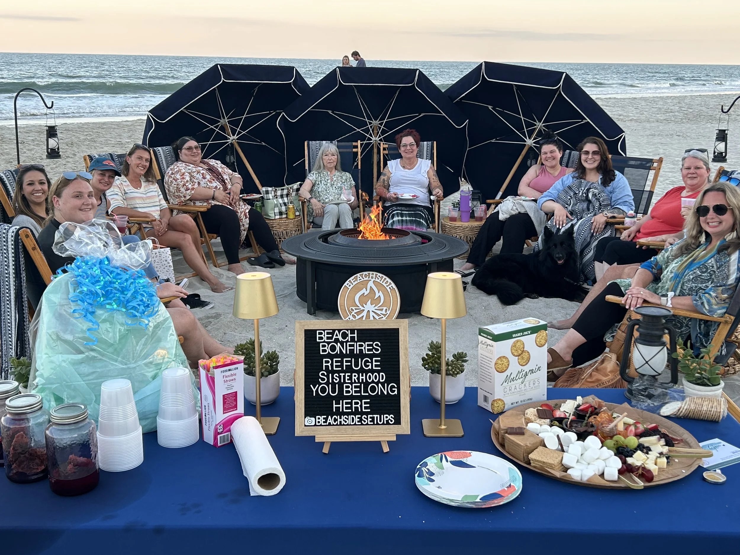 Beachside bonfire setup with a group of women sitting around a fire pit on the beach, surrounded by umbrellas, snacks, and drinks.