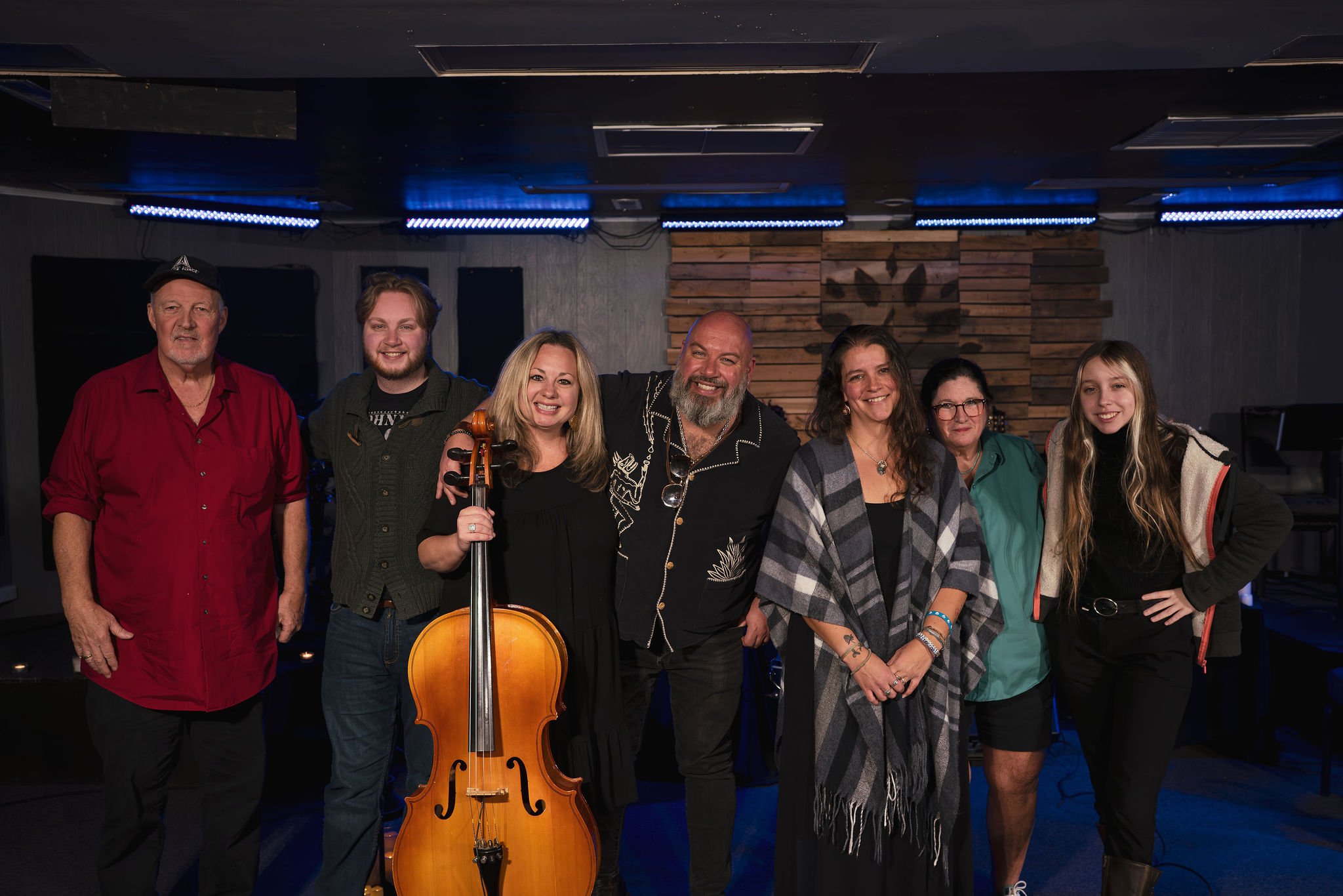 Group of eight people standing in a music venue, with one holding a cello, smiling at the camera, in front of a wooden wall decorated with a butterfly image and blue stage lights.