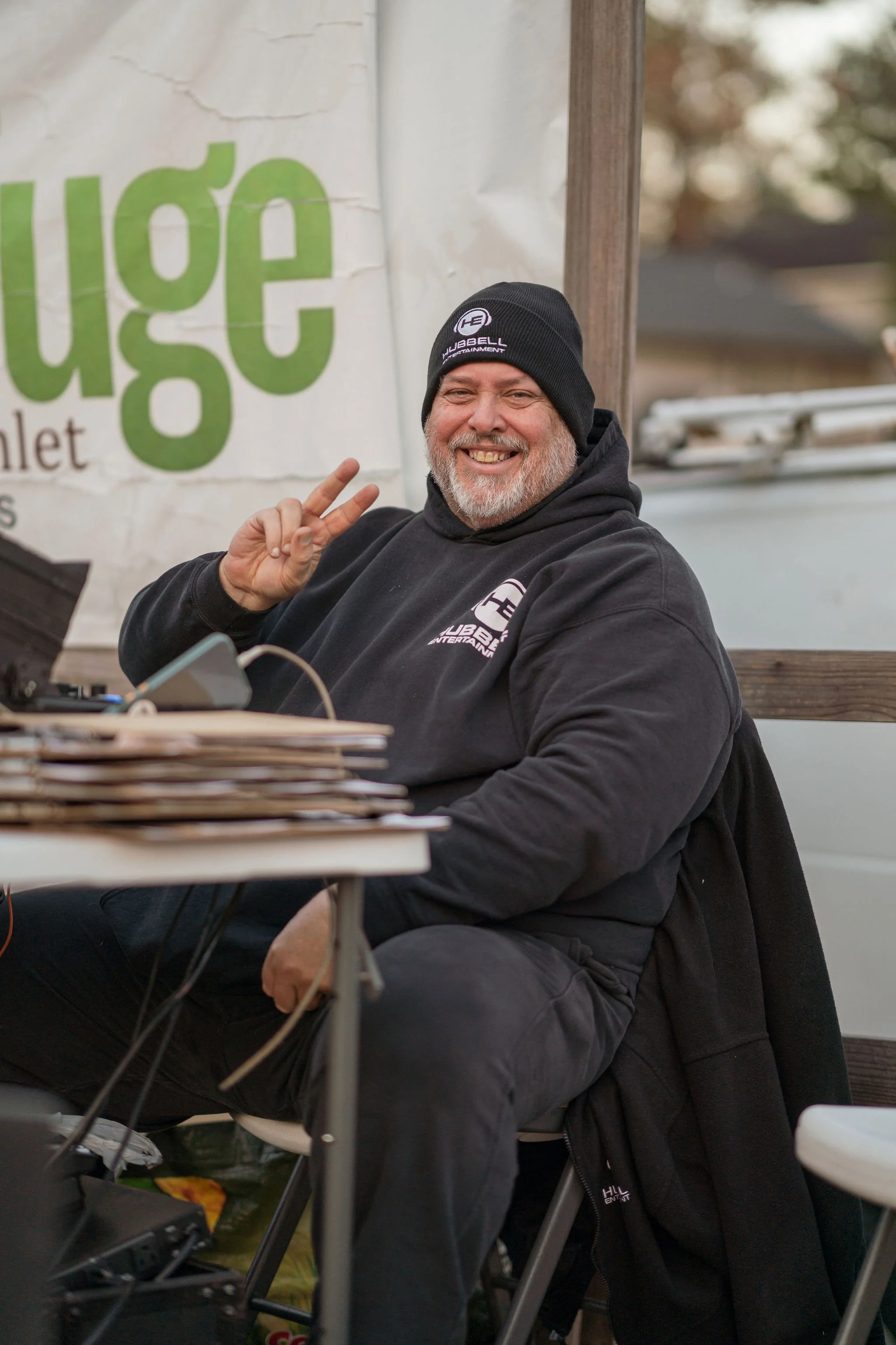 A smiling man with a gray beard and mustache wearing a black beanie and black hoodie, making a peace sign with his right hand, sitting outdoors at a table with computer equipment. There is a large sign with green letters partially visible behind him.