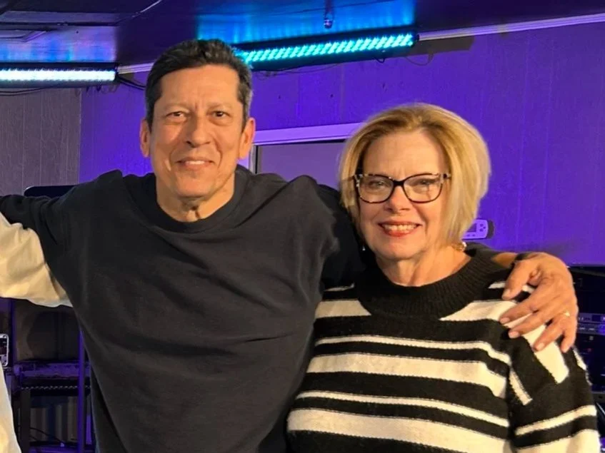 A man and a woman standing close together indoors with purple and blue lighting, smiling at the camera. The man is wearing a black long-sleeve shirt, and the woman is wearing glasses and a black and white striped sweater.