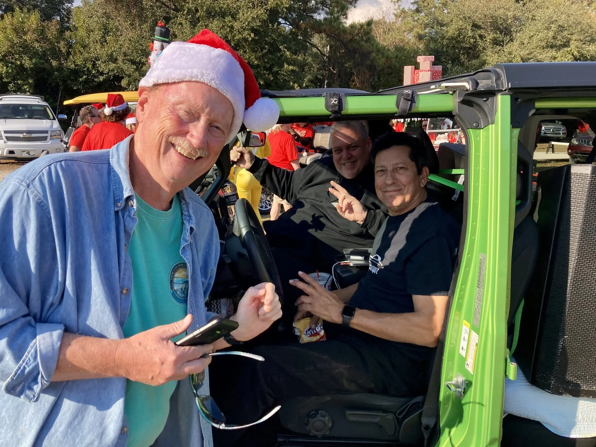 A group of people, including a man wearing a Santa hat, gathered at an outdoor event with golf carts and cars in the background. The people are smiling and appear to be enjoying a festive celebration.