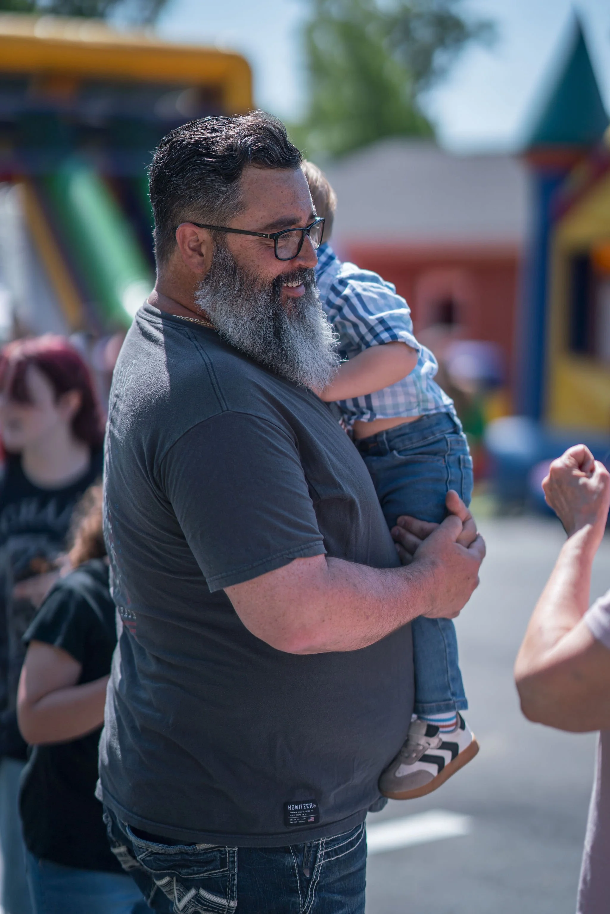 An older man with glasses and a beard is holding a young boy. The setting appears to be outdoors at a fair or amusement park, with rides visible in the background. The man and boy are smiling and interacting with someone outside the frame.