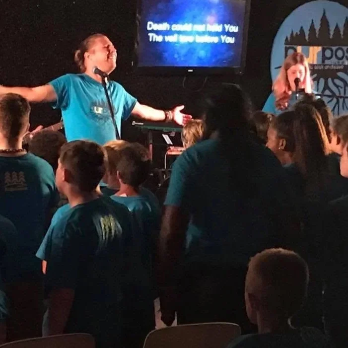 A speaker with outstretched arms addressing a group of young people in a dark room. A woman is singing or speaking into a microphone, with a screen behind displaying text. The audience, mostly young individuals wearing matching teal shirts, are listening and facing the speaker.