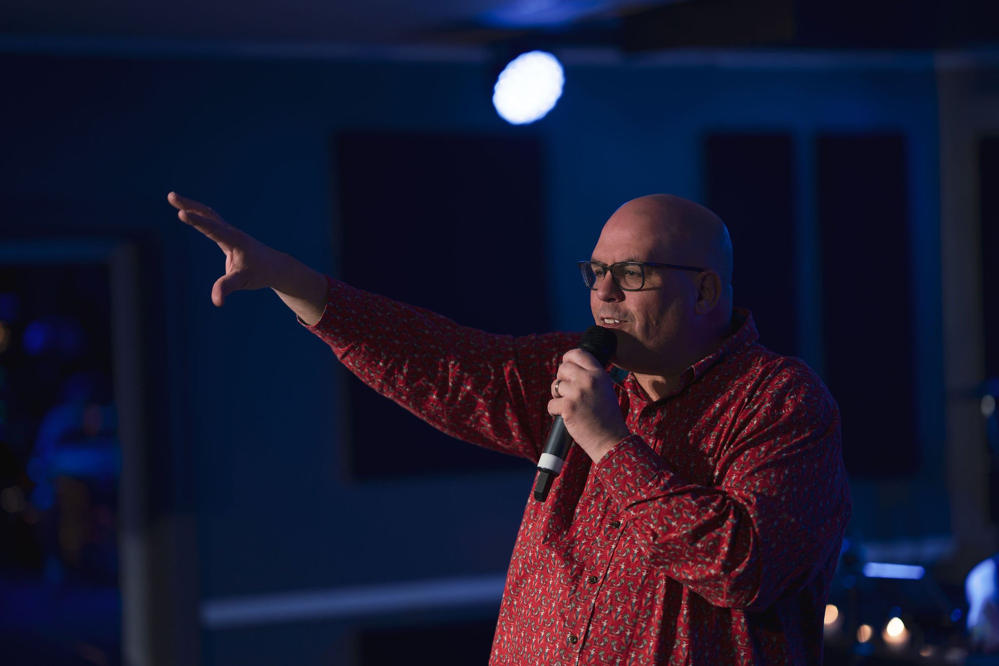 A bald man with glasses wearing a red patterned shirt is speaking into a microphone and gesturing with his right hand, in a dark indoor setting with blue lighting.