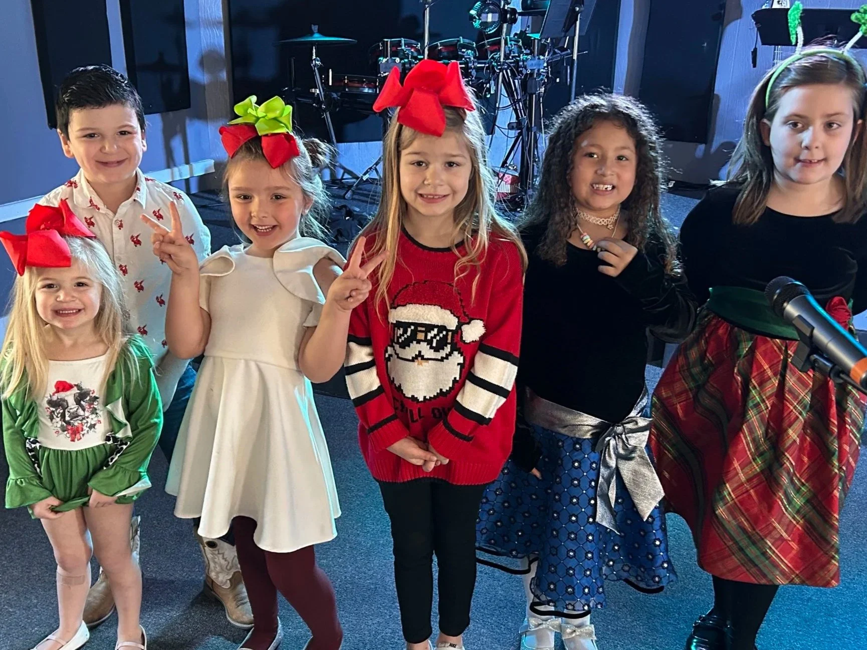 Group of children at a festive event, some making peace signs, dressed in holiday-themed outfits, with musical instruments in the background.