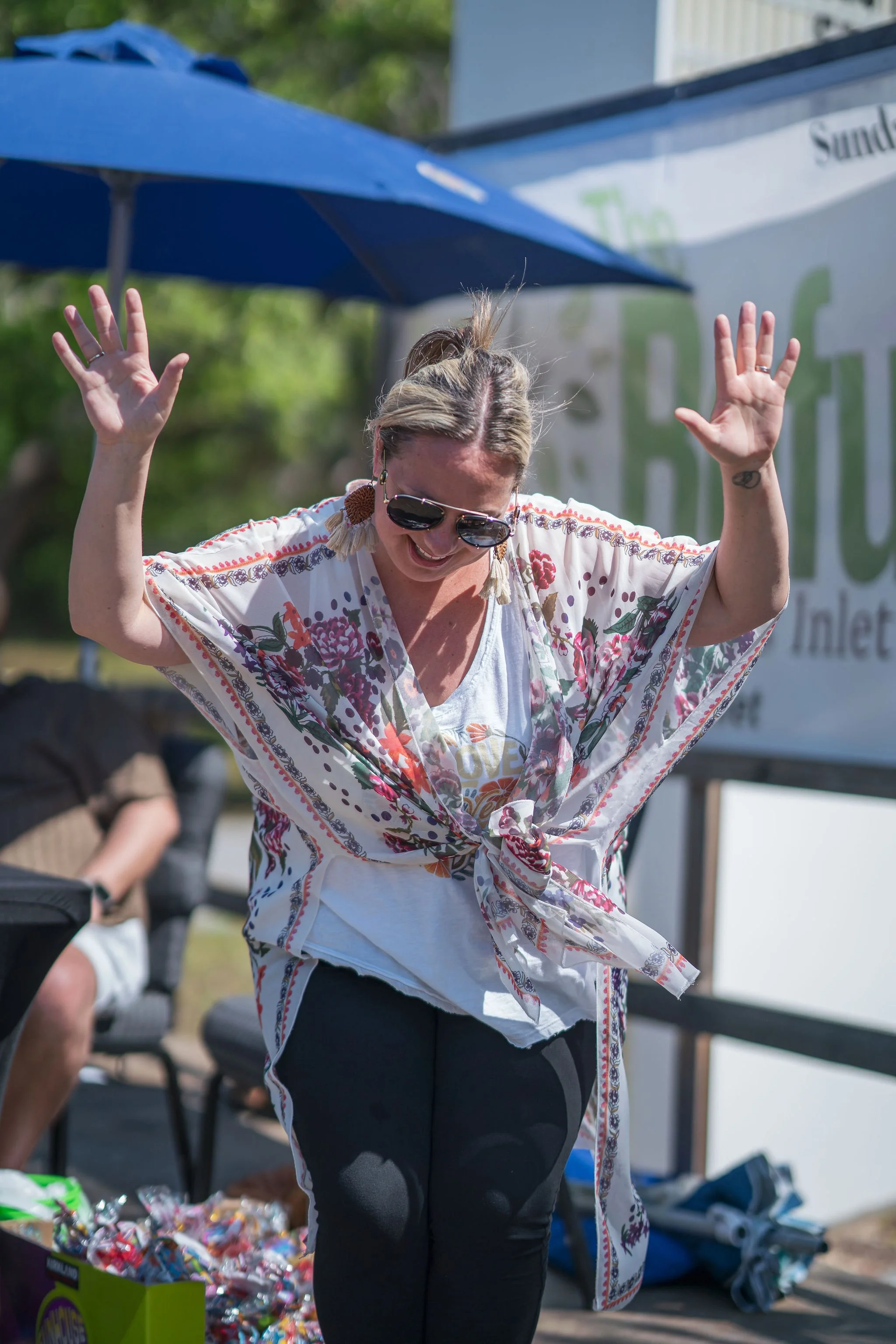 Woman wearing sunglasses with her hands raised, smiling outdoors with a table of candy and a blue umbrella nearby.