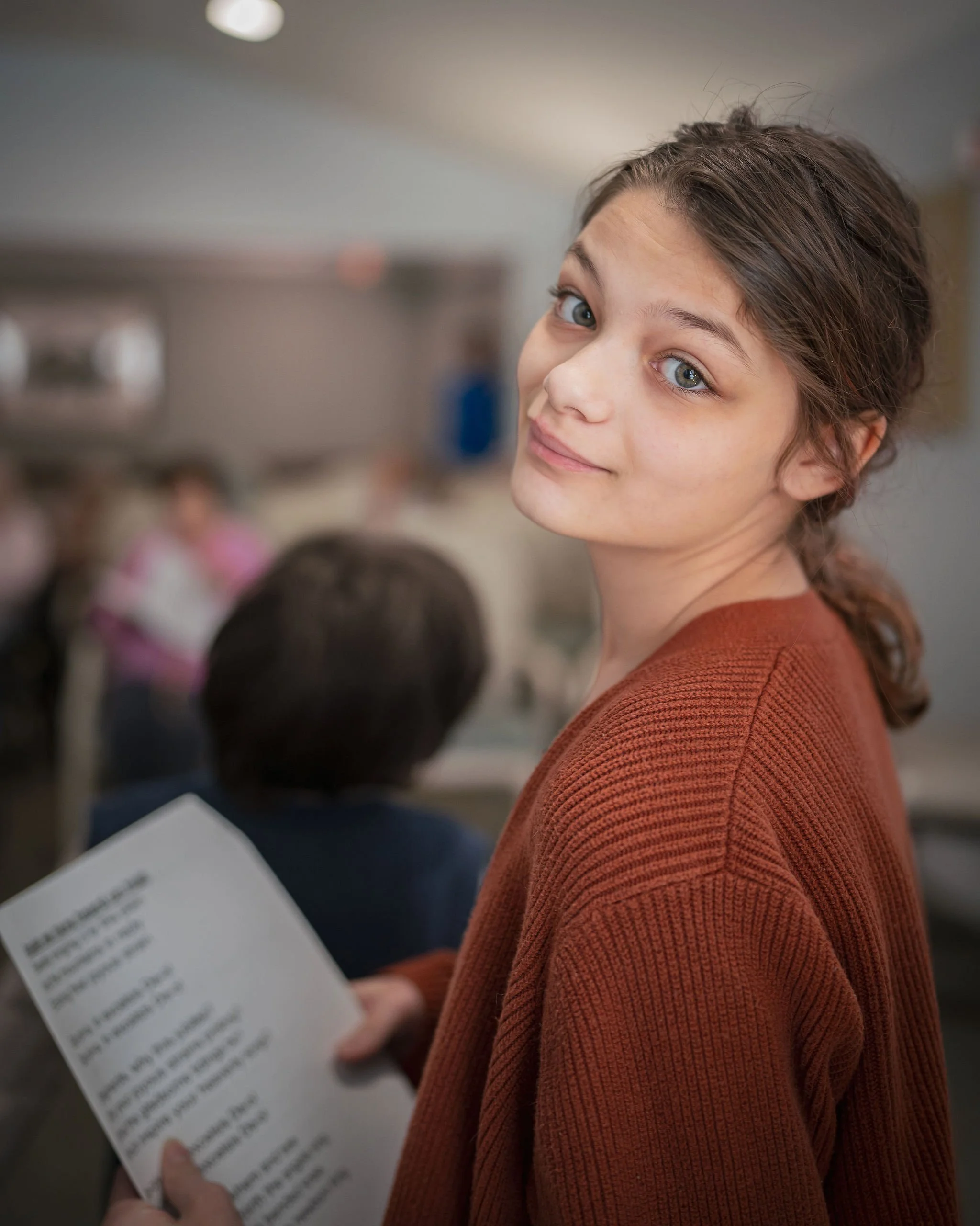 A young woman with brown hair and blue eyes looking over her shoulder at the camera while holding a piece of paper, in a room with other people in the background.