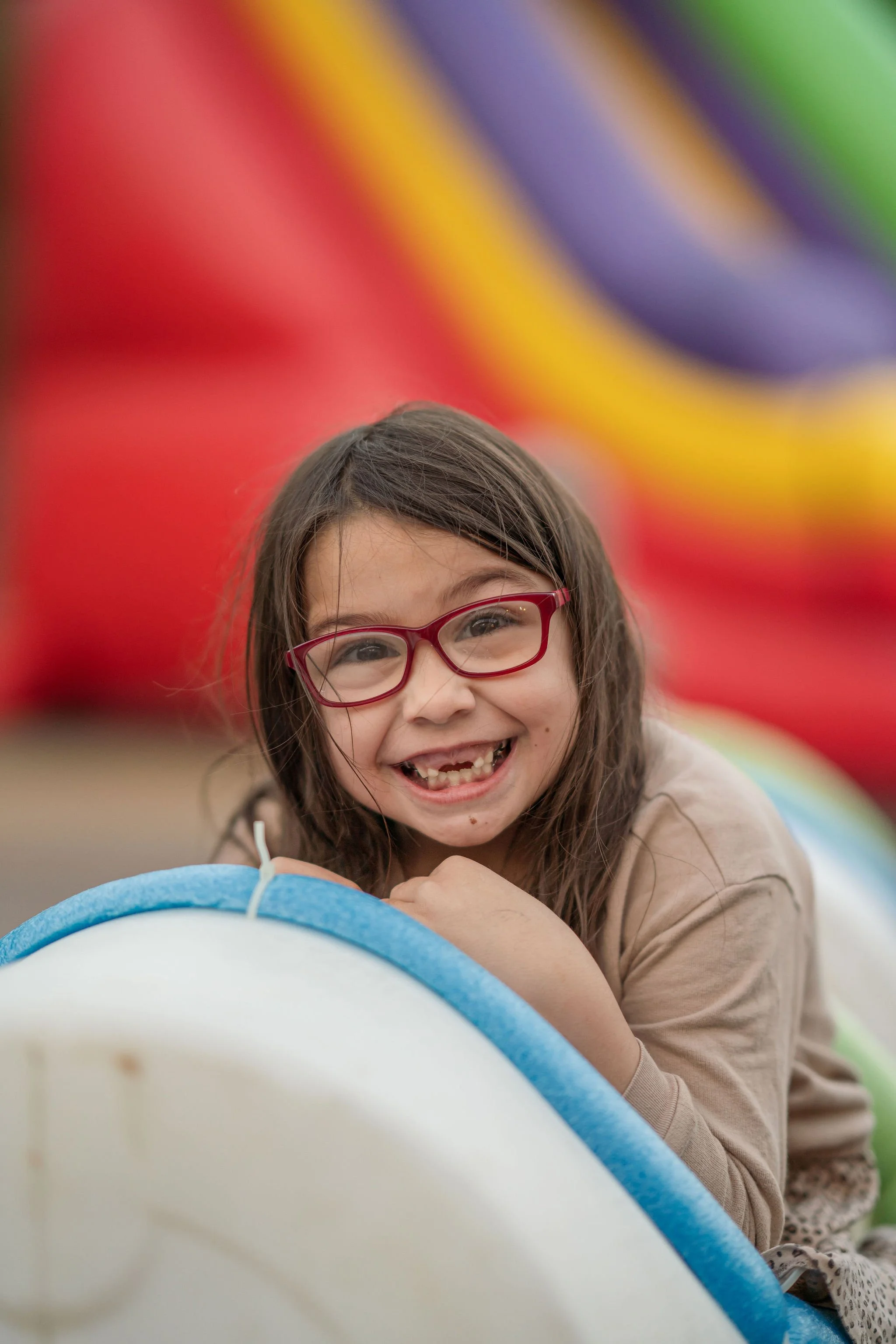 A young girl with long brown hair, wearing glasses, smiling while playing on an inflatable bouncy house with colorful background.