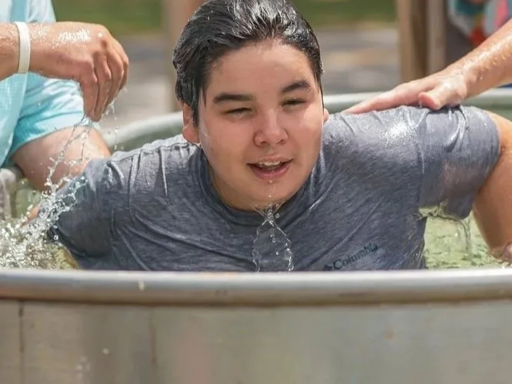 A young man in a gray t-shirt being baptized outdoors in a metal tub with water, with other hands supporting him.