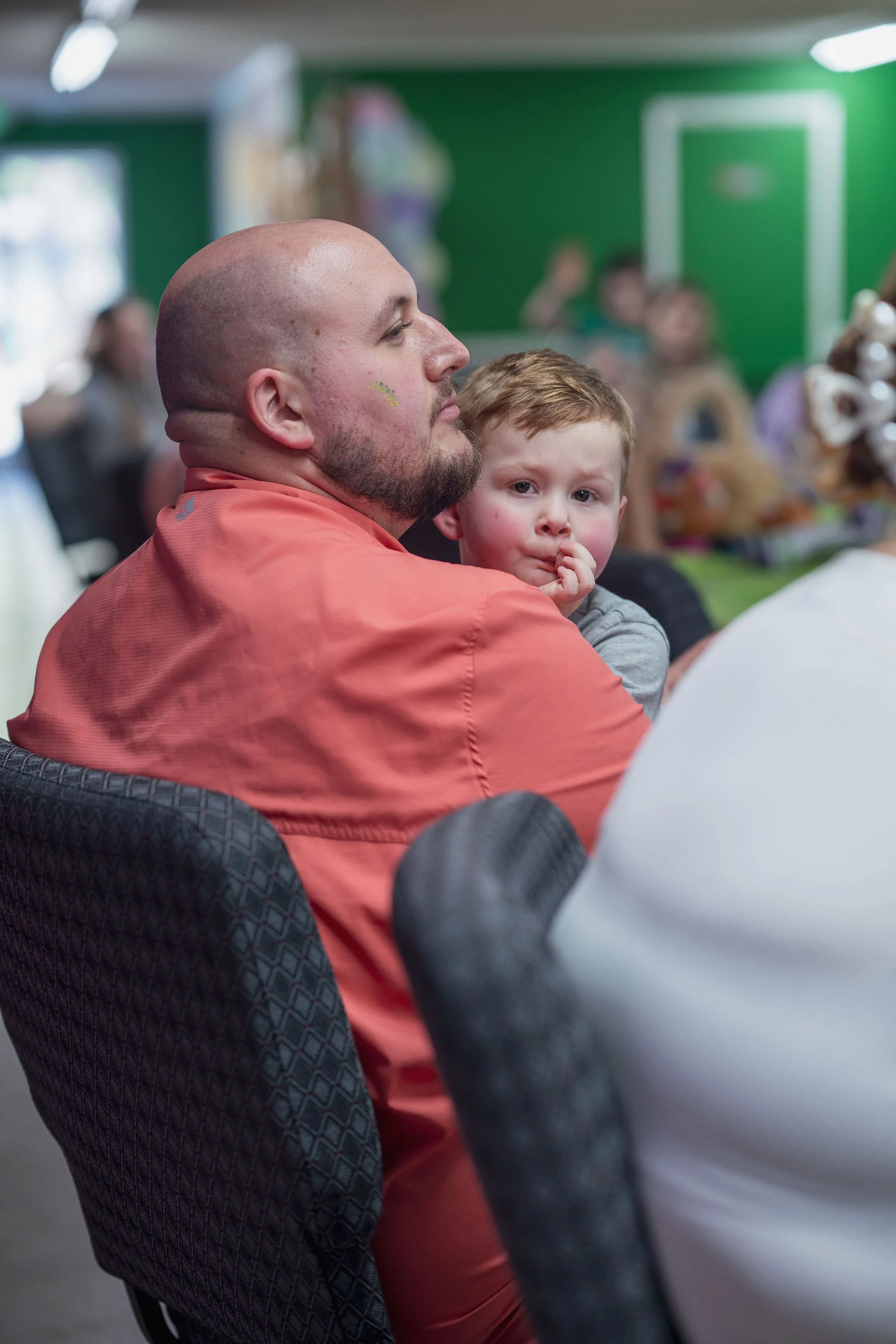 A man with a beard and a close-shaven head, wearing an orange shirt, sitting on a dark patterned chair, and a young boy with light brown hair, wearing a gray shirt, sitting on the man's lap. They are in a room with a bright green wall, and there are 