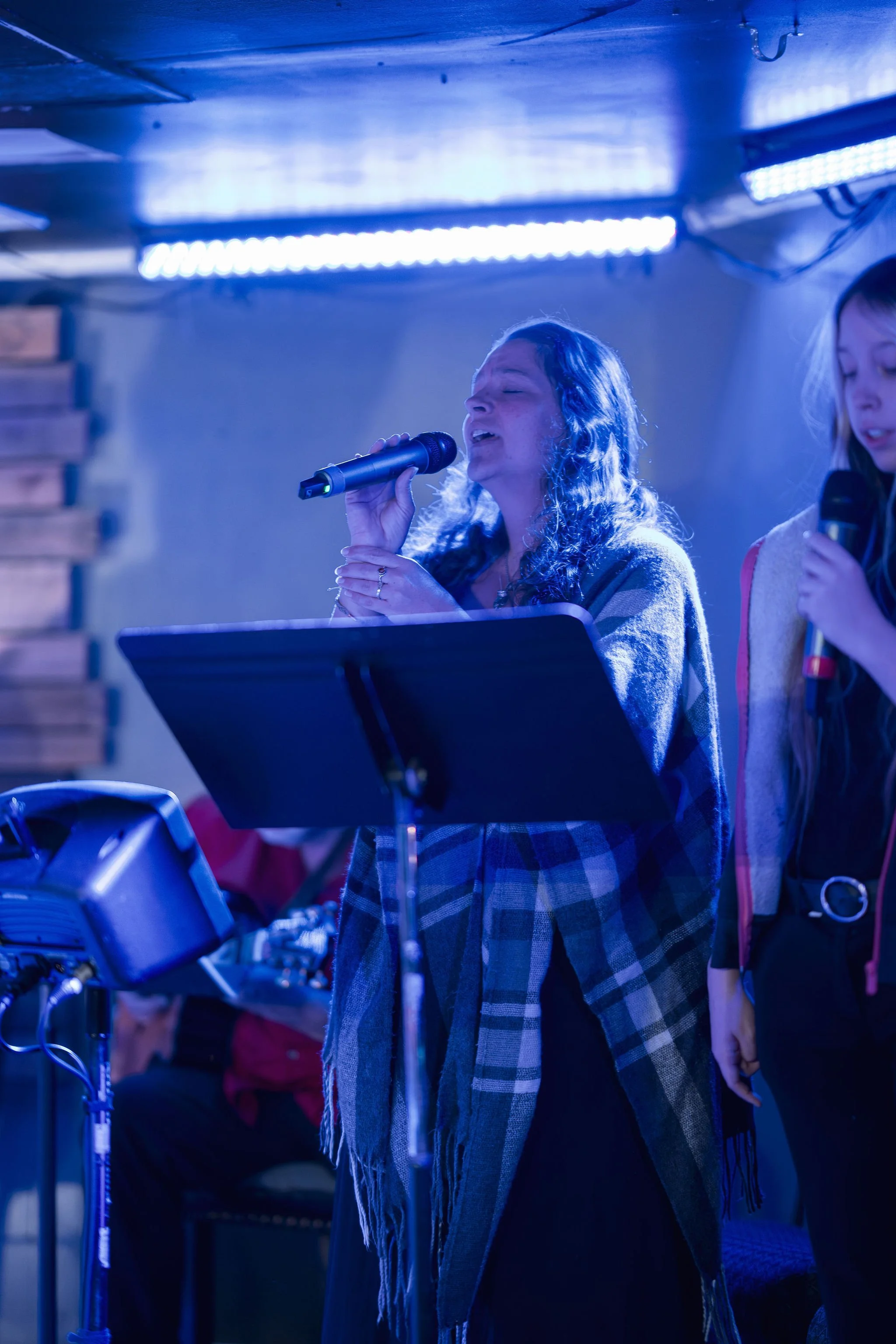 A woman singing passionately into a microphone while wearing a plaid coat, with another person partially visible next to her, under blue stage lighting.