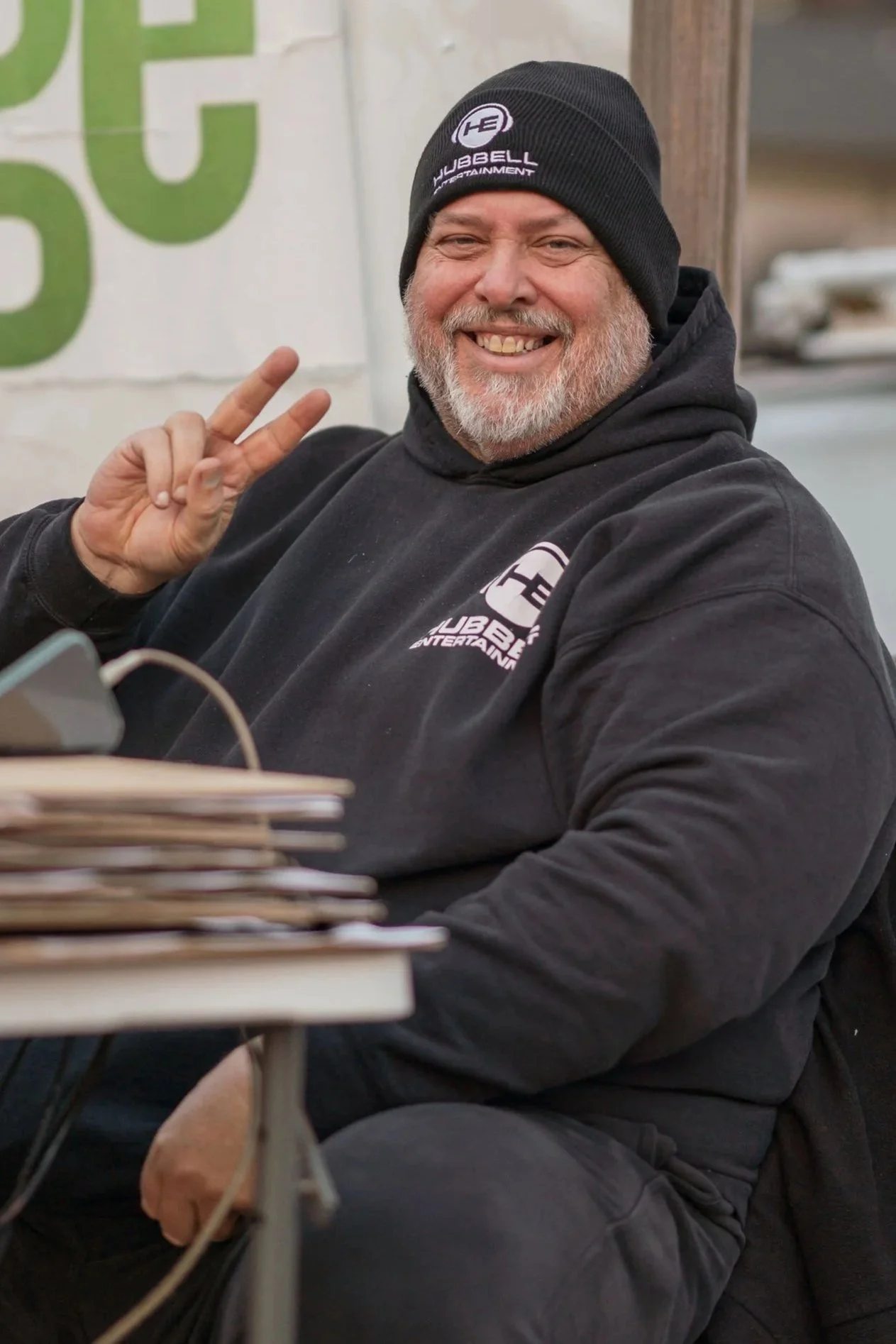 A smiling man wearing a black hoodie and a black beanie with the logo 'Hubbel Entertainment' making a peace sign gesture. He is sitting at a table with a stack of magazines or newspapers in front of him.