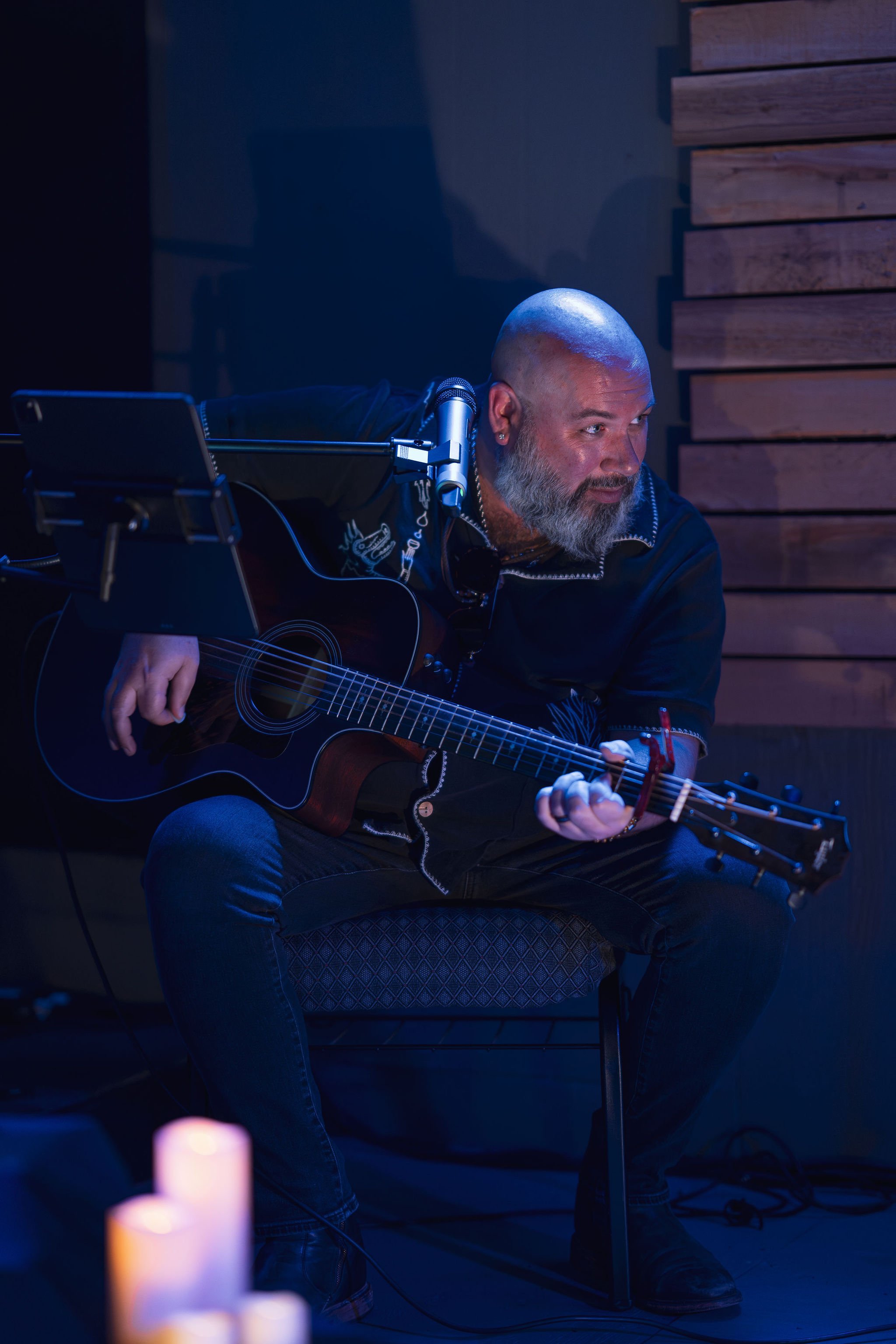A man with a shaved head and a beard playing an acoustic guitar, seated in a dimly lit room with wooden wall panels behind him. There is a microphone positioned near his face.