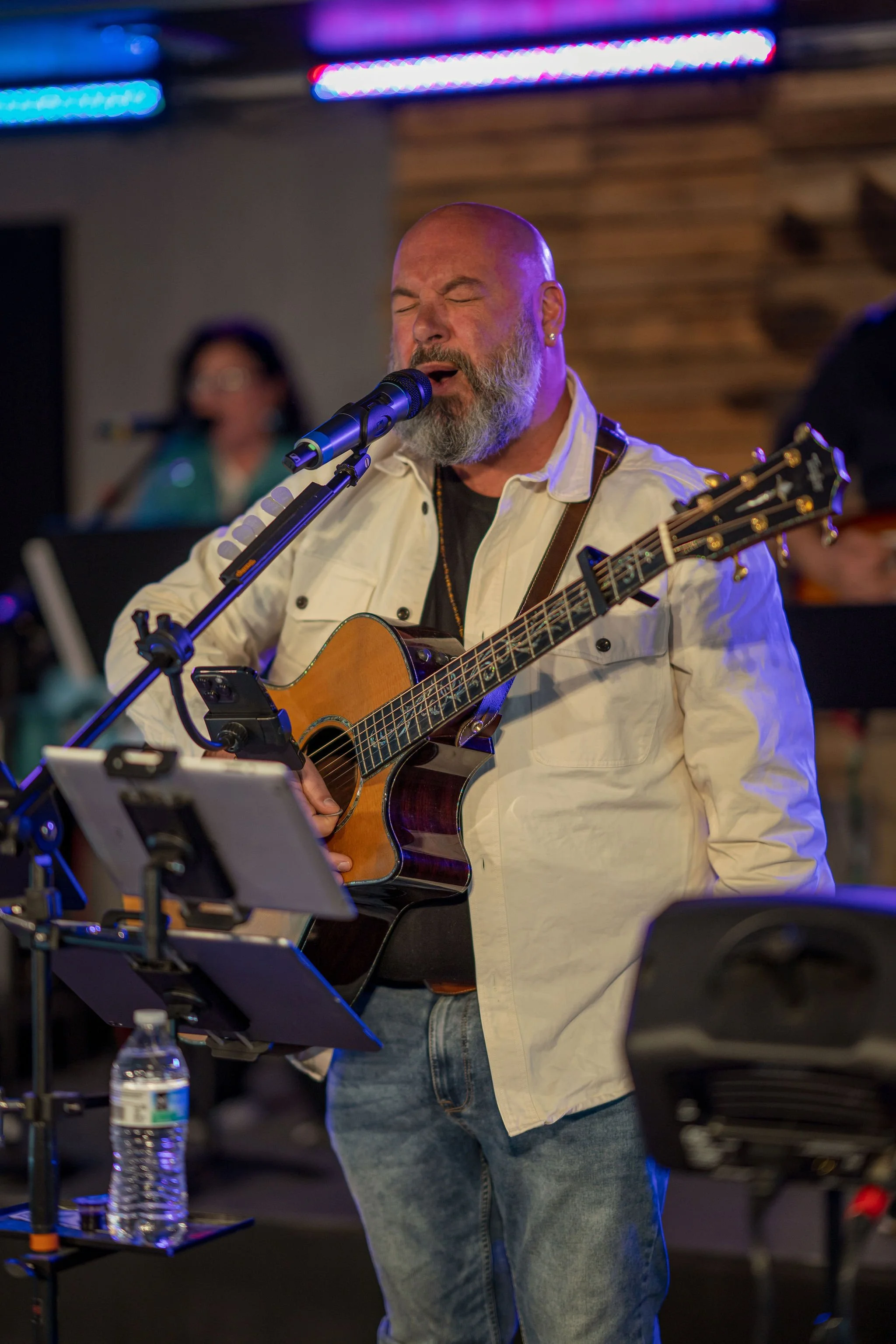 A man with a beard and earrings passionately singing into a microphone while playing an acoustic guitar during a live performance. There are blue and purple stage lights and a woman in the background.