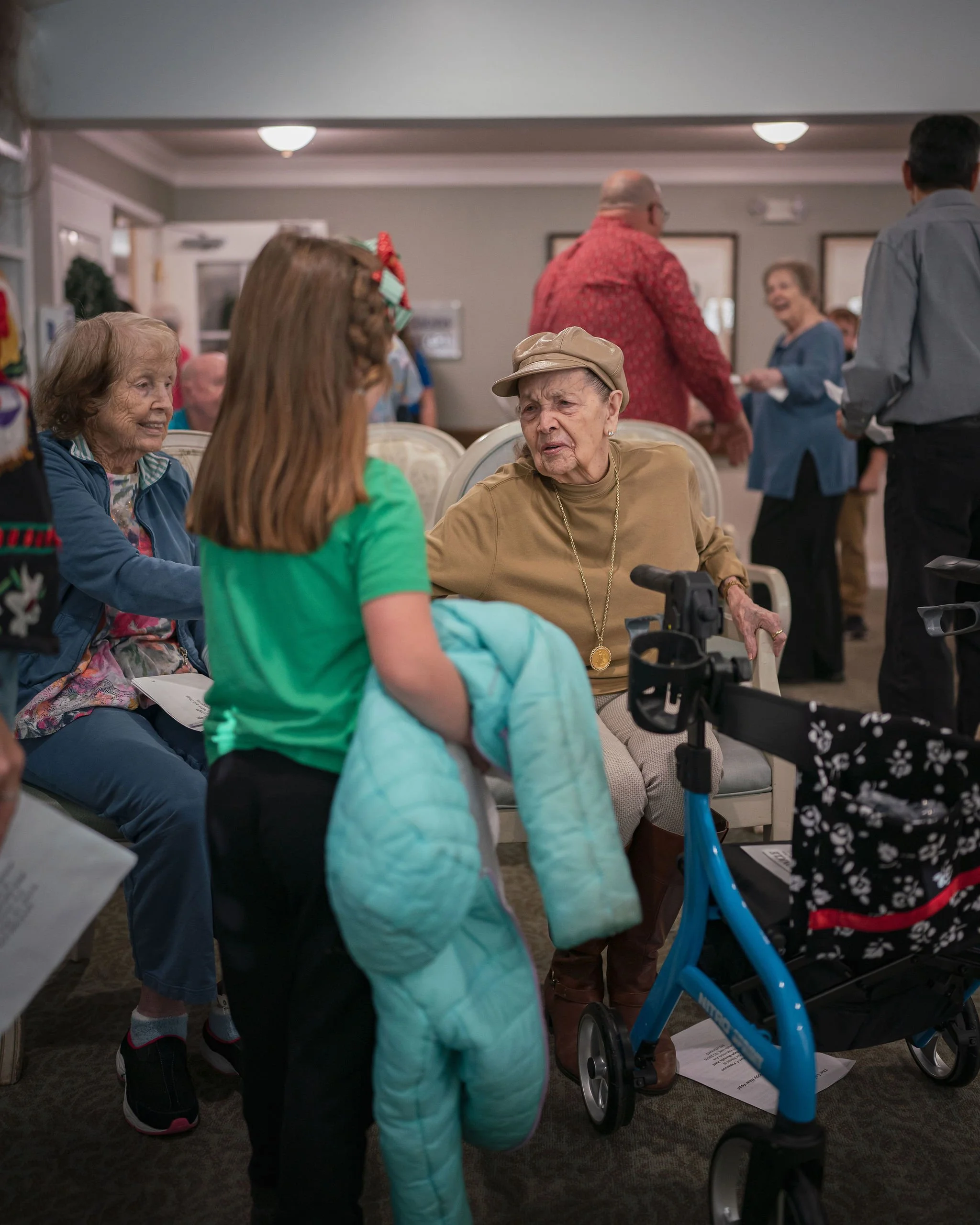 People socializing in a community center or senior living facility, with elderly women seated and engaging in conversation, and others standing or walking around, including a woman in a green shirt and a woman with a beige hat in the foreground.