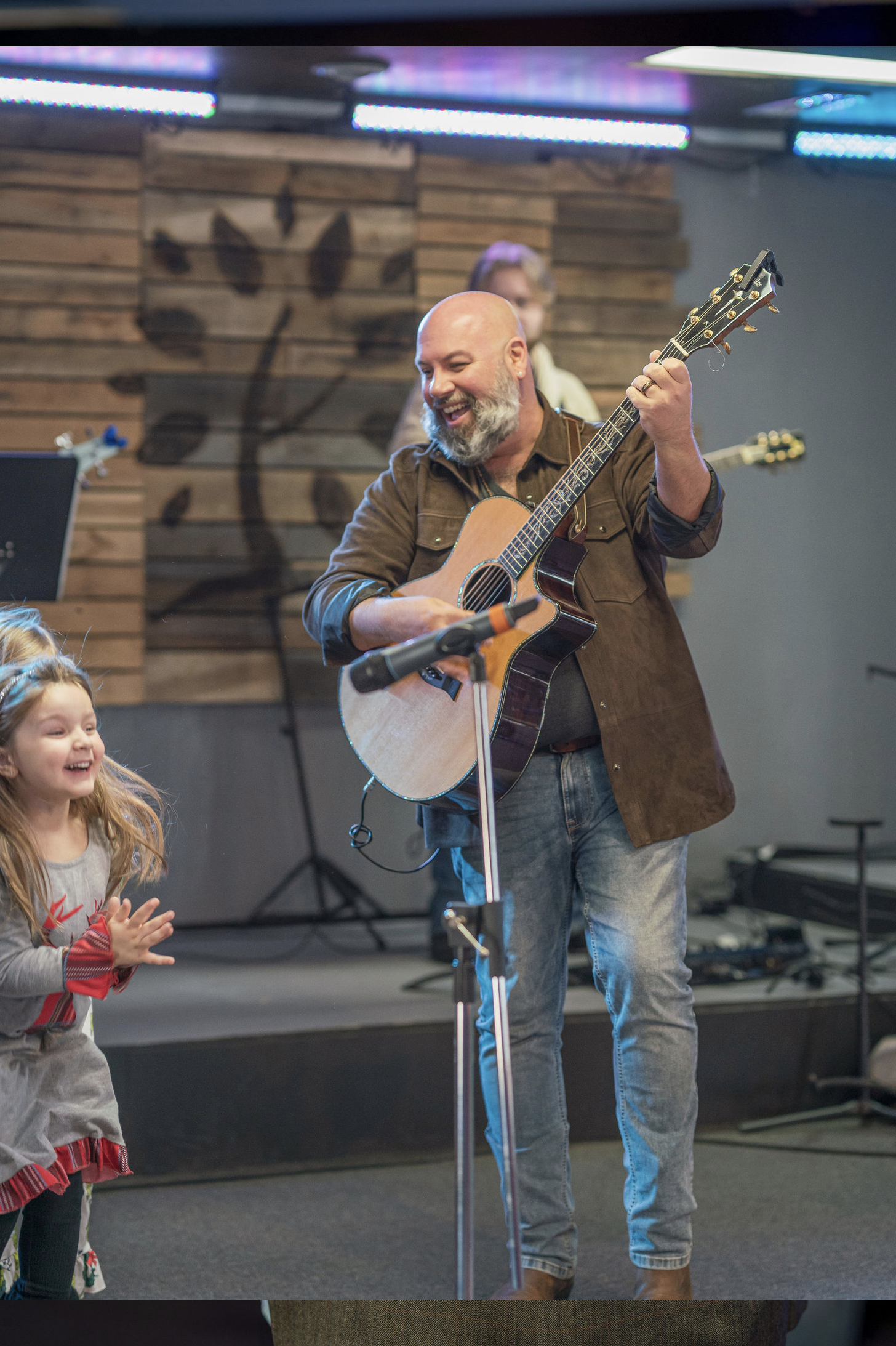 Man playing an acoustic guitar smiling while children clap and cheer in front of him at an indoor event with wooden wall art and stage lighting.