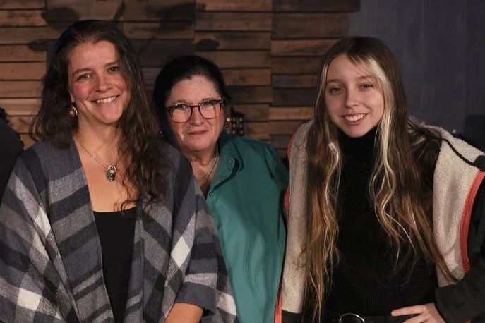 Three women standing close together and smiling in front of a wooden wall. The woman on the left has long curly hair and wears a plaid shawl. The woman in the middle has dark hair, glasses, and is wearing a teal jacket. The young woman on the right has long blonde hair and is dressed in a black top with a beige jacket.