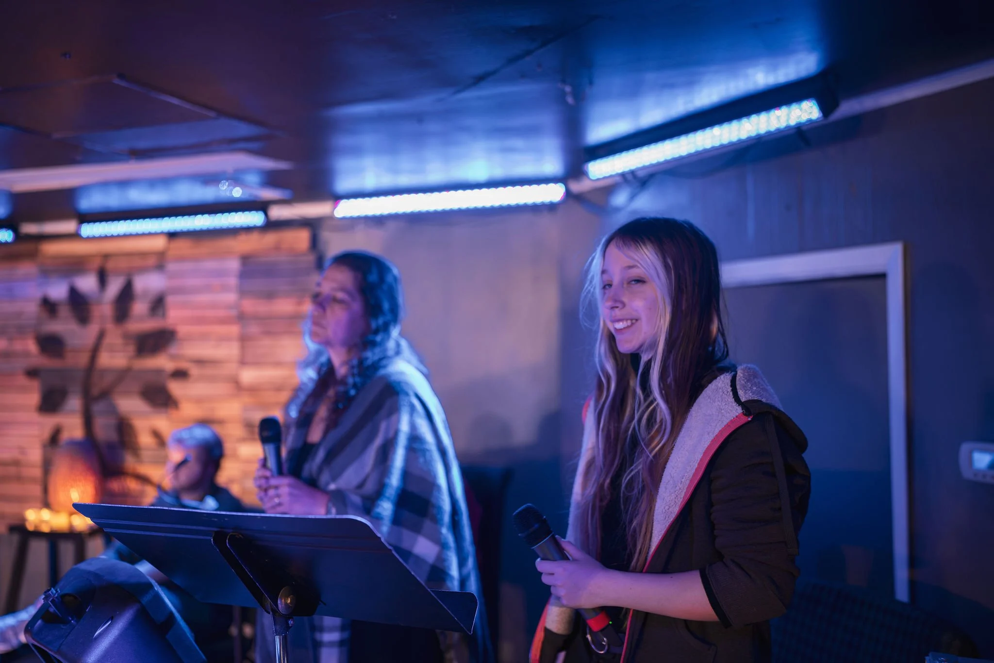 Two women standing in front of microphones in a dimly lit room with blue and purple lighting, one of them smiling, likely at a karaoke or music event.