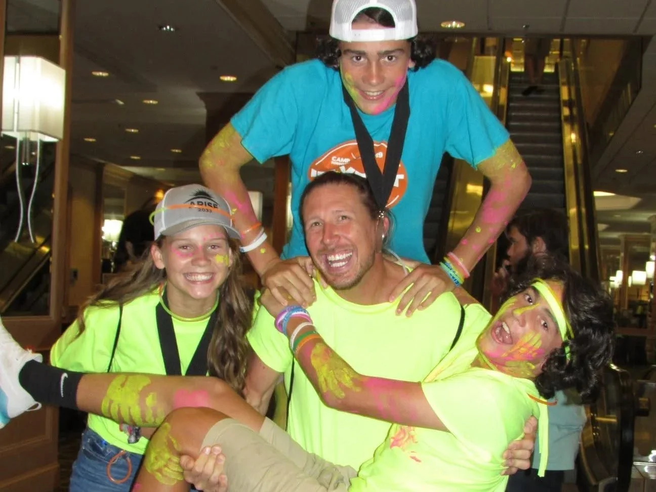 Group of four young people celebrating at a colorful event, with paint splatters on their faces and clothing, surrounded by indoor hotel lobby decor.