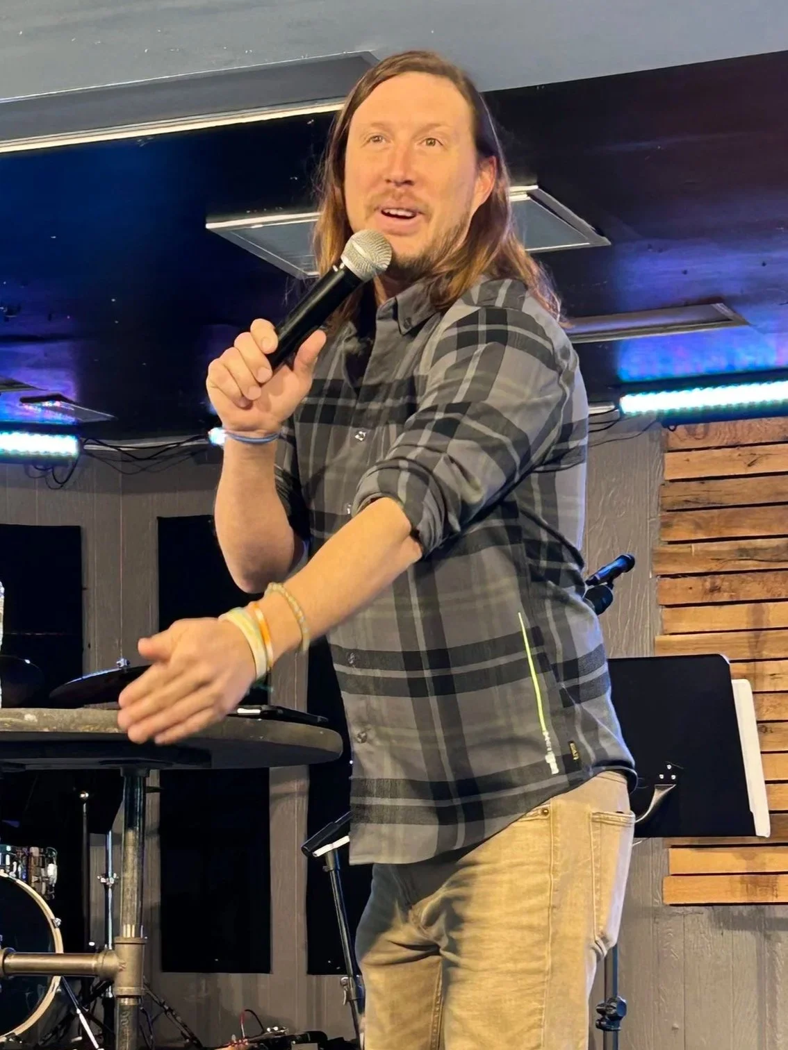 Man performing stand-up comedy on stage with microphone in hand, wearing a plaid shirt and beige pants, in a venue with wood and black paneling decor.