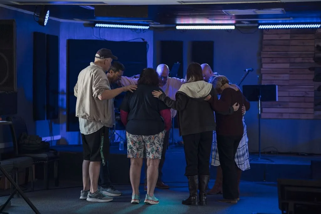 A group of seven people stand in a circle with their heads bowed and hands on each other's shoulders in a prayer circle in a dimly lit room with musical instruments and stage lighting.