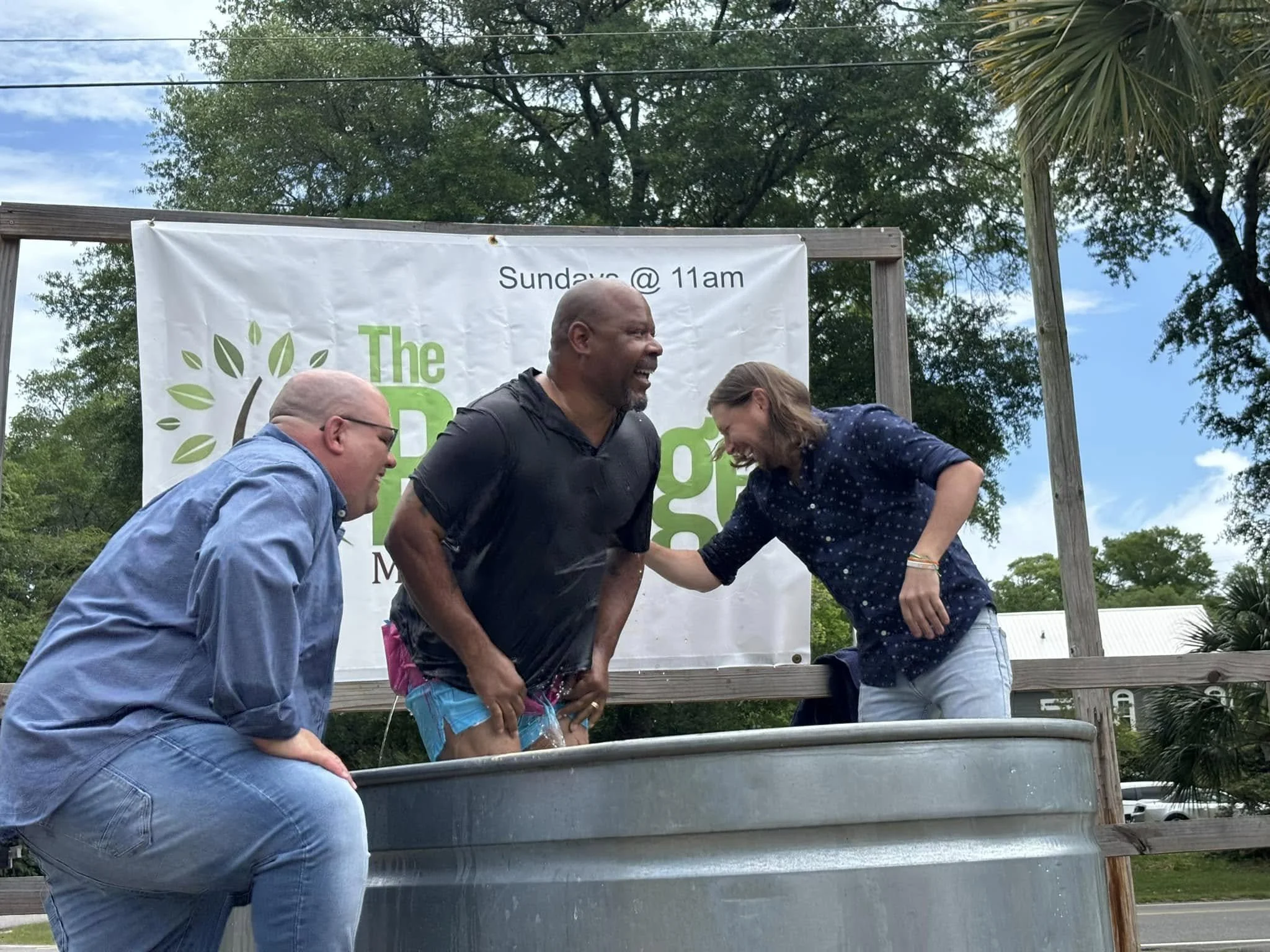 Group of four people, three men and one woman, standing outside in front of a banner that reads 'The' and 'Sunday @ 11am'. They appear to be joyful, engaging with each other near a metal baptismal pool, with trees and a blue sky in the background.