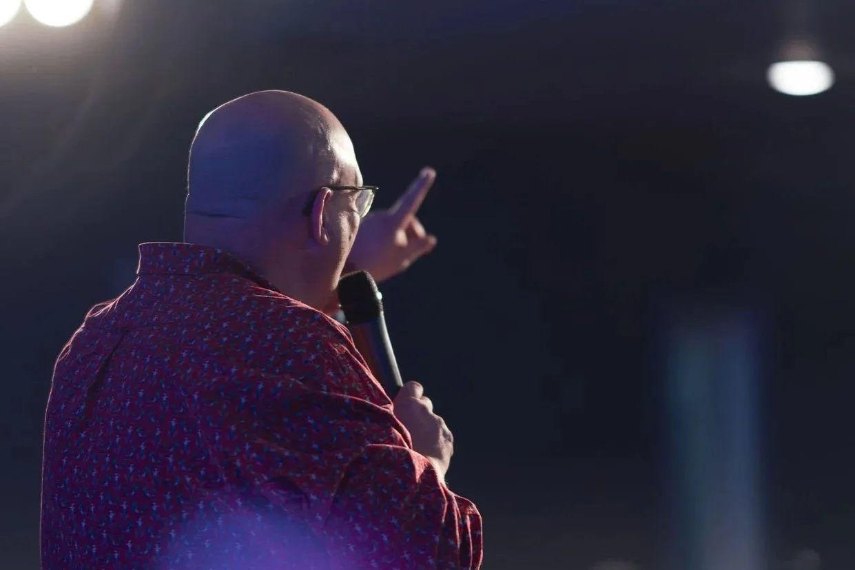 A bald man wearing glasses, a gray blazer, and a blue shirt, holding a microphone and speaking with his eyes closed. He is standing in front of a blurred background with people and colorful lights.