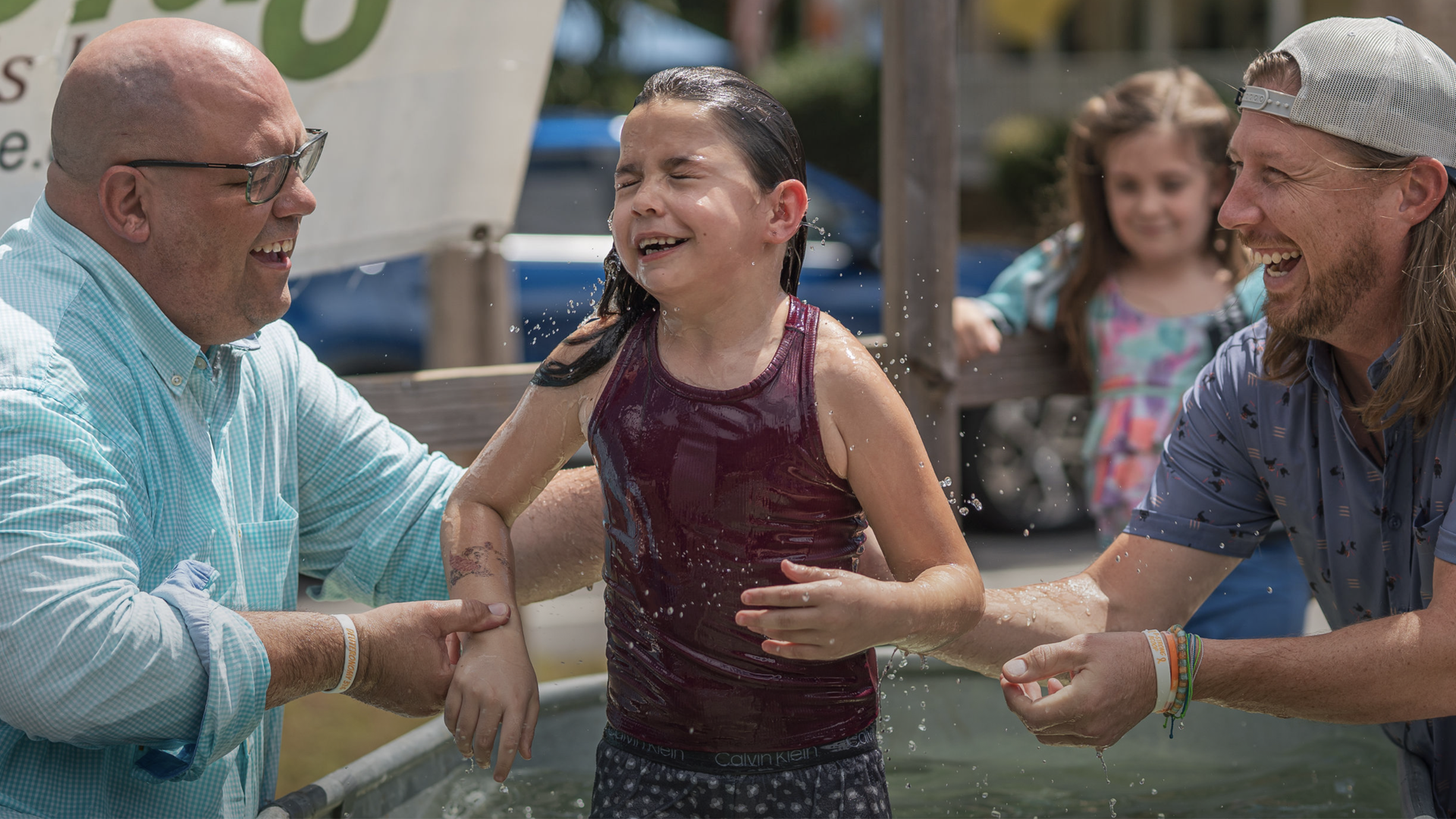 A young girl in a maroon tank top getting baptized in a shallow pool, smiling with eyes closed, with two men holding her and a girl in the background watching.