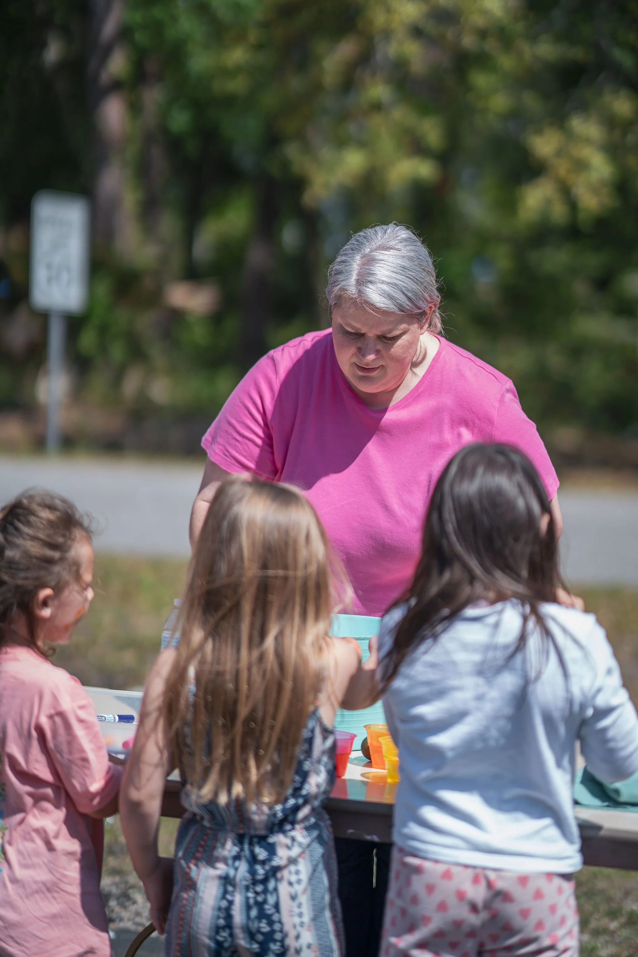 A woman with gray hair wearing a pink shirt is surrounded by children and appears to be looking at something or talking. The children have their backs to the camera. They are outdoors with trees and a street sign visible in the background.