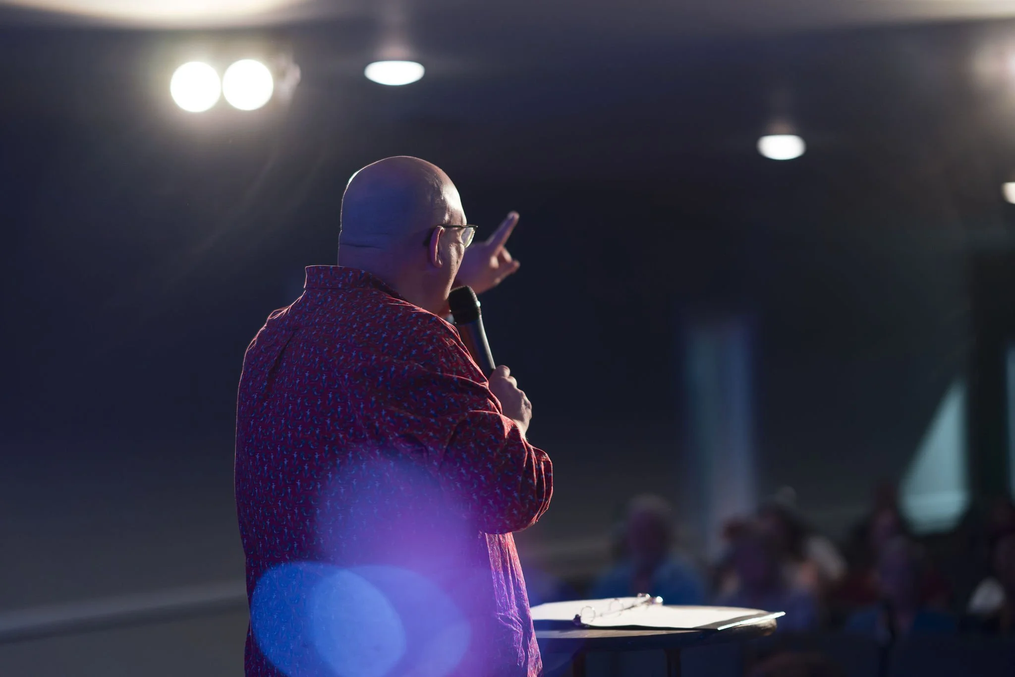 A bald man wearing glasses and a patterned red shirt is speaking into a microphone on stage, gesturing with his right hand, with a blurred audience in the background and bright stage lights overhead.