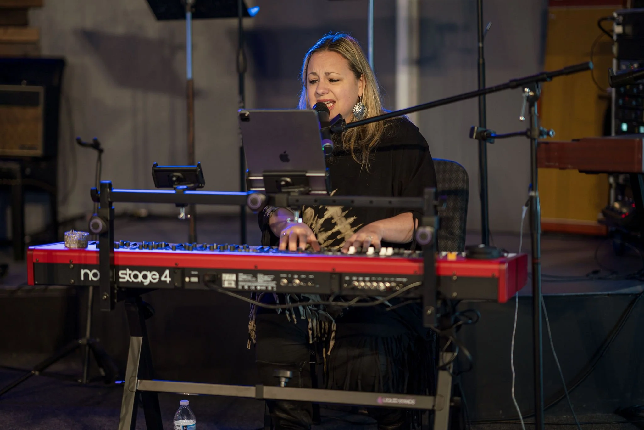 A woman singing into a microphone while playing a keyboard on stage with music equipment and stands around her.