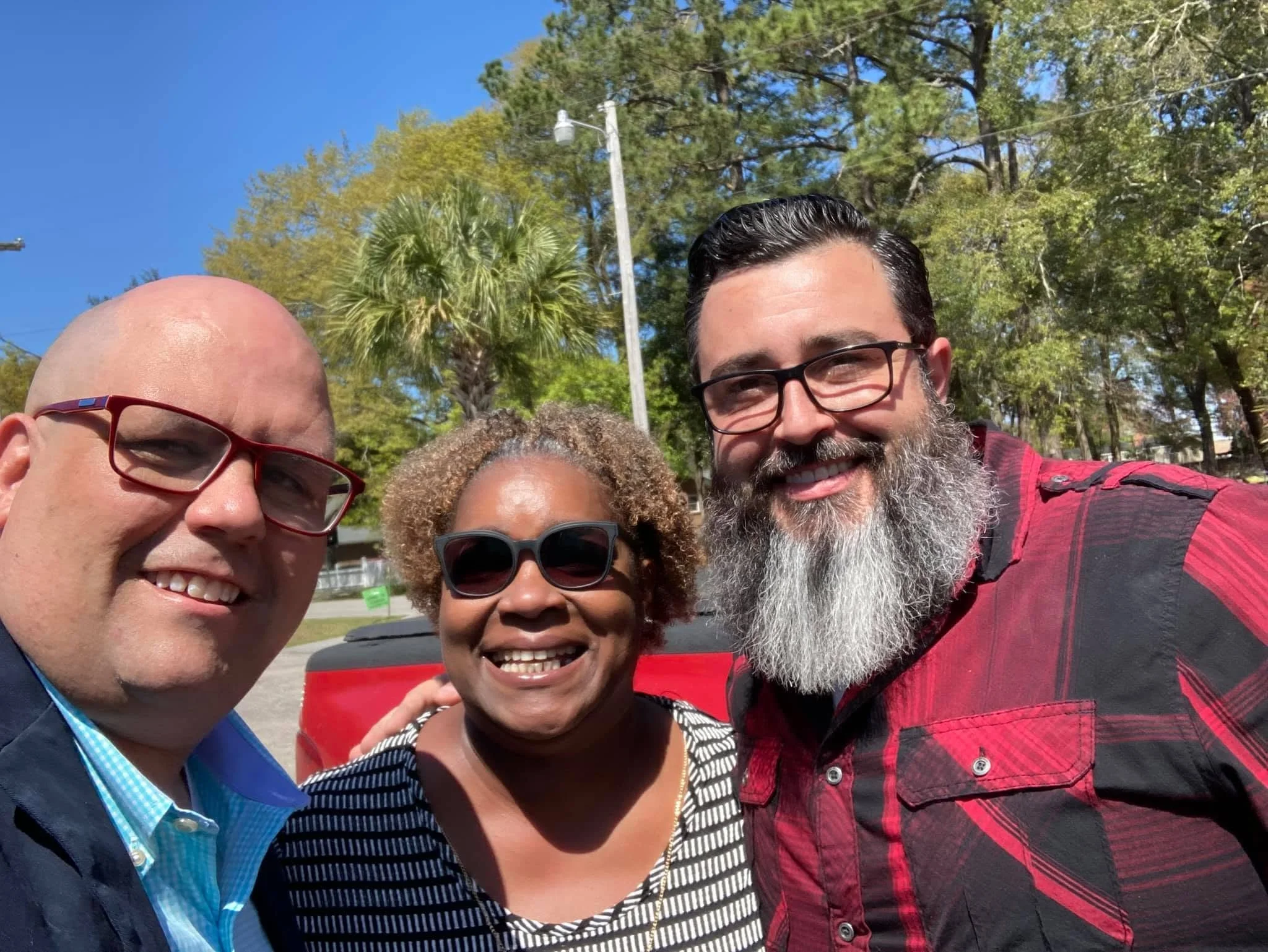 A group of four diverse people smiling outdoors on a sunny day. Trees, a utility pole, and a clear blue sky are in the background.