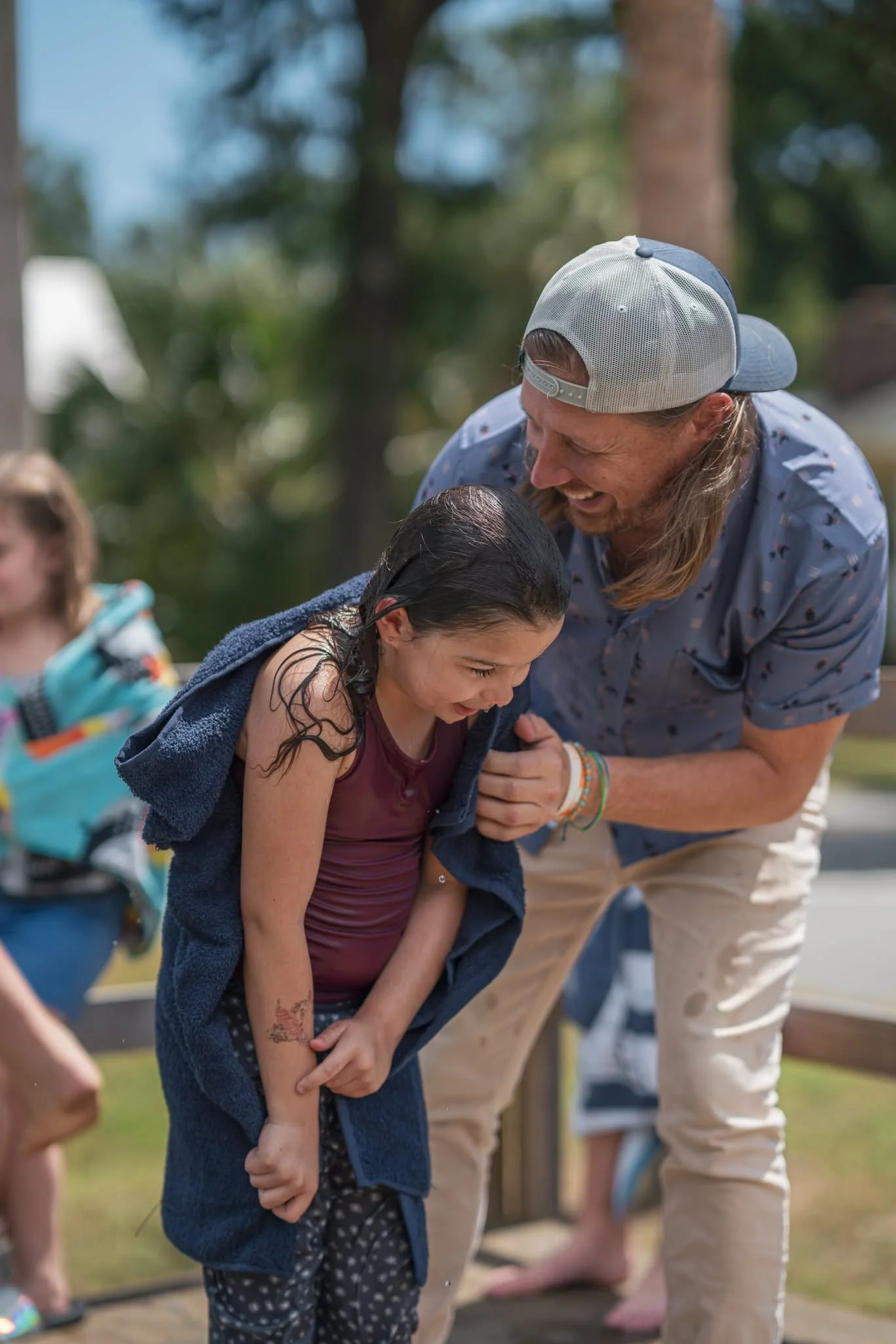 A man with long hair and a cap leaning over and holding a young girl with a towel over her shoulders, at an outdoor event.