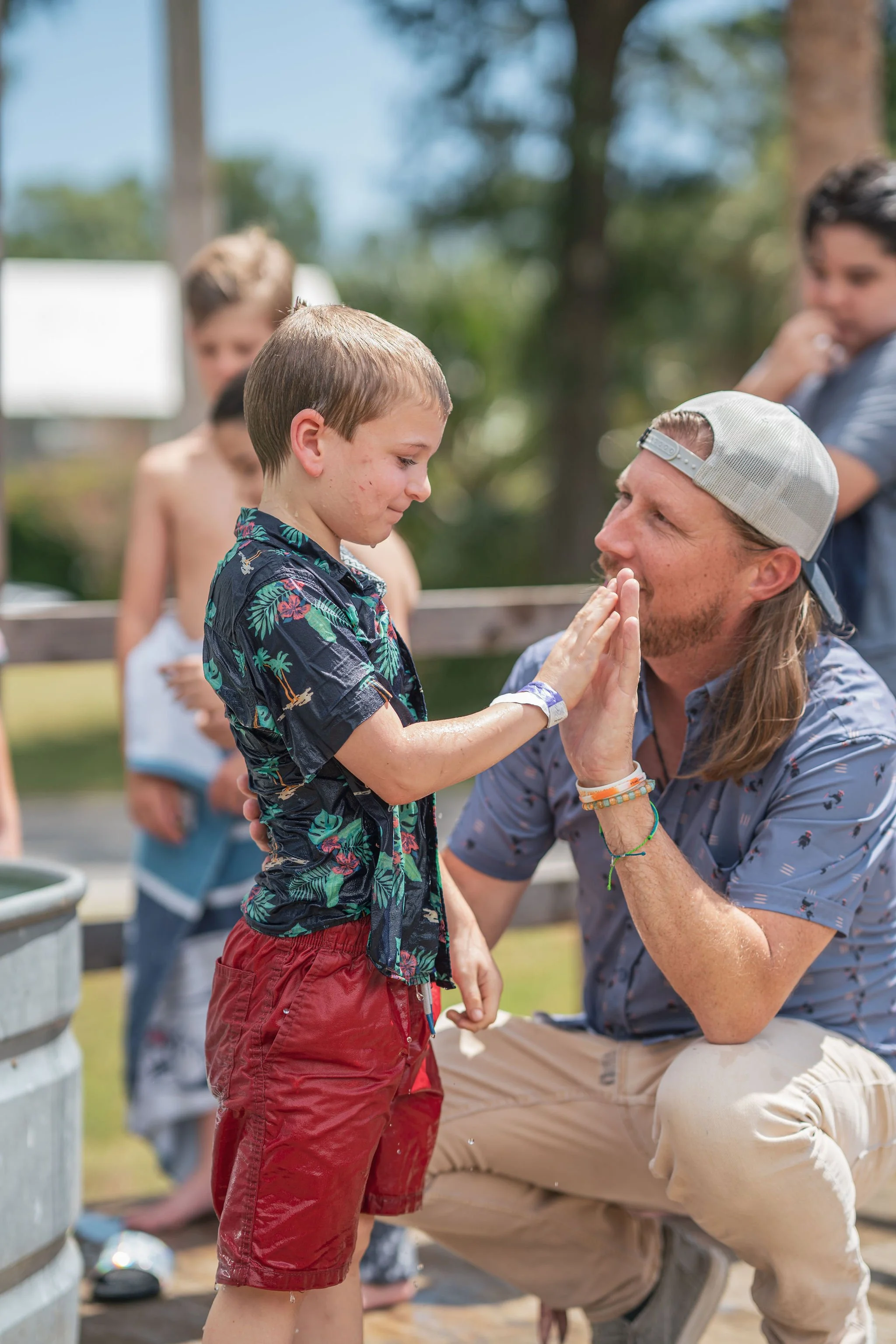 A young boy is giving a high-five to a man wearing a baseball cap outdoors, with a line of children in swimwear behind them. The setting appears to be a summer outdoor activity.