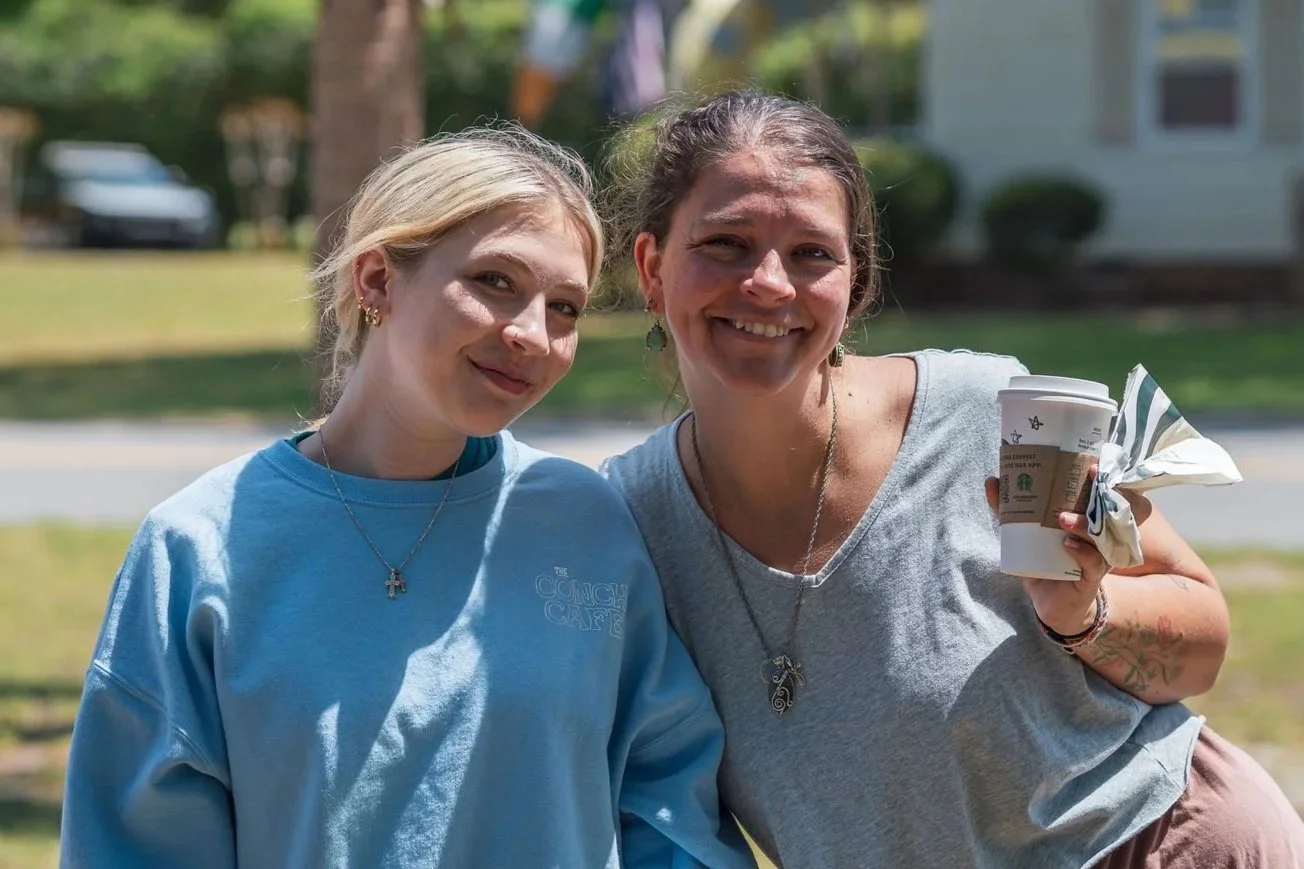 Two smiling women standing outdoors, one holding a Starbucks coffee cup wrapped with a napkin. The woman on the left has blonde hair, is wearing a light blue sweatshirt and a necklace. The woman on the right has dark hair, is wearing a grey t-shirt, and has tattoos on her arm.