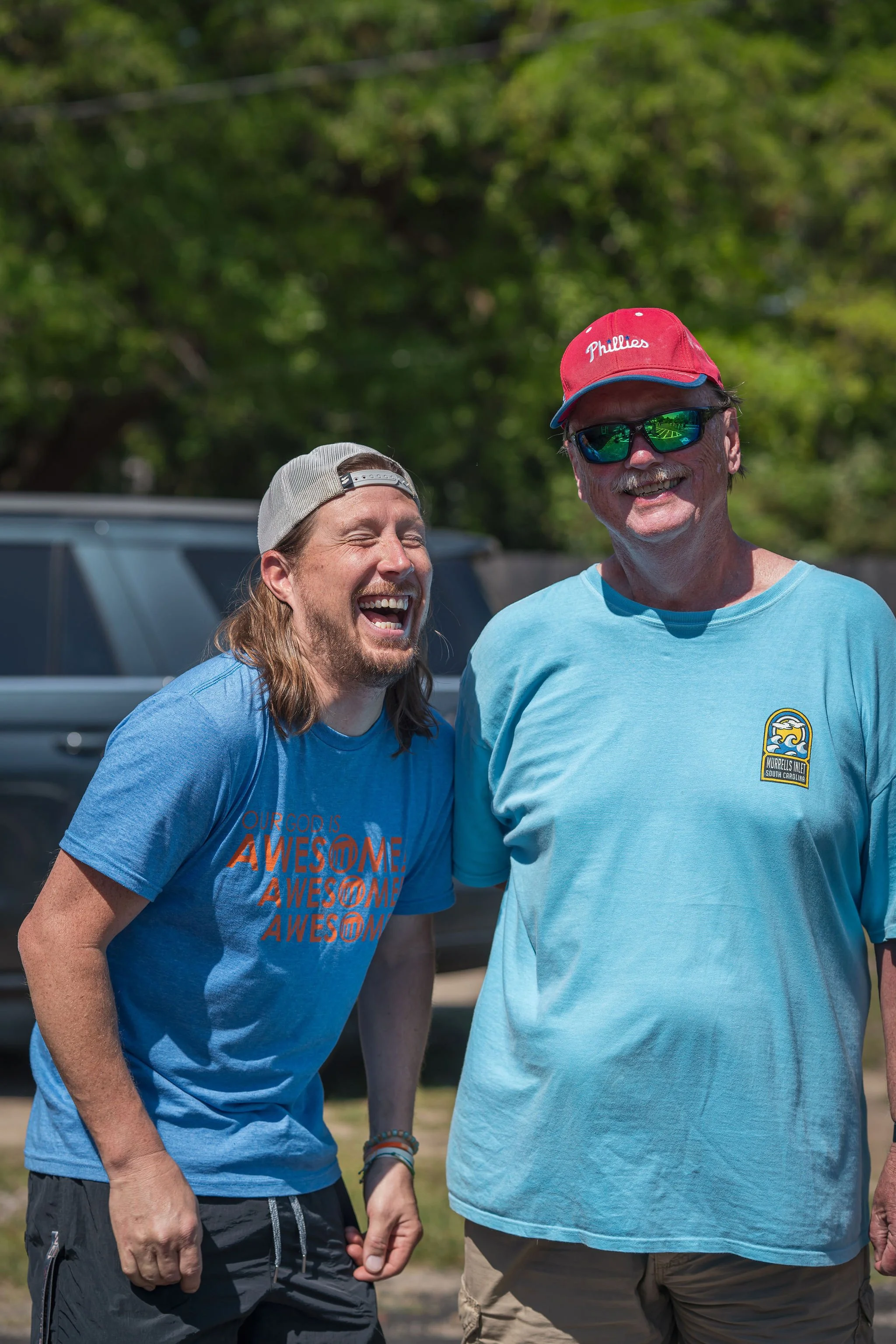 Two men smiling outdoors, one wearing a gray cap backward and a blue t-shirt with orange writing, the other wearing a red cap with 'Phillies' embroidered in white, dark sunglasses, and a light blue t-shirt with a logo.