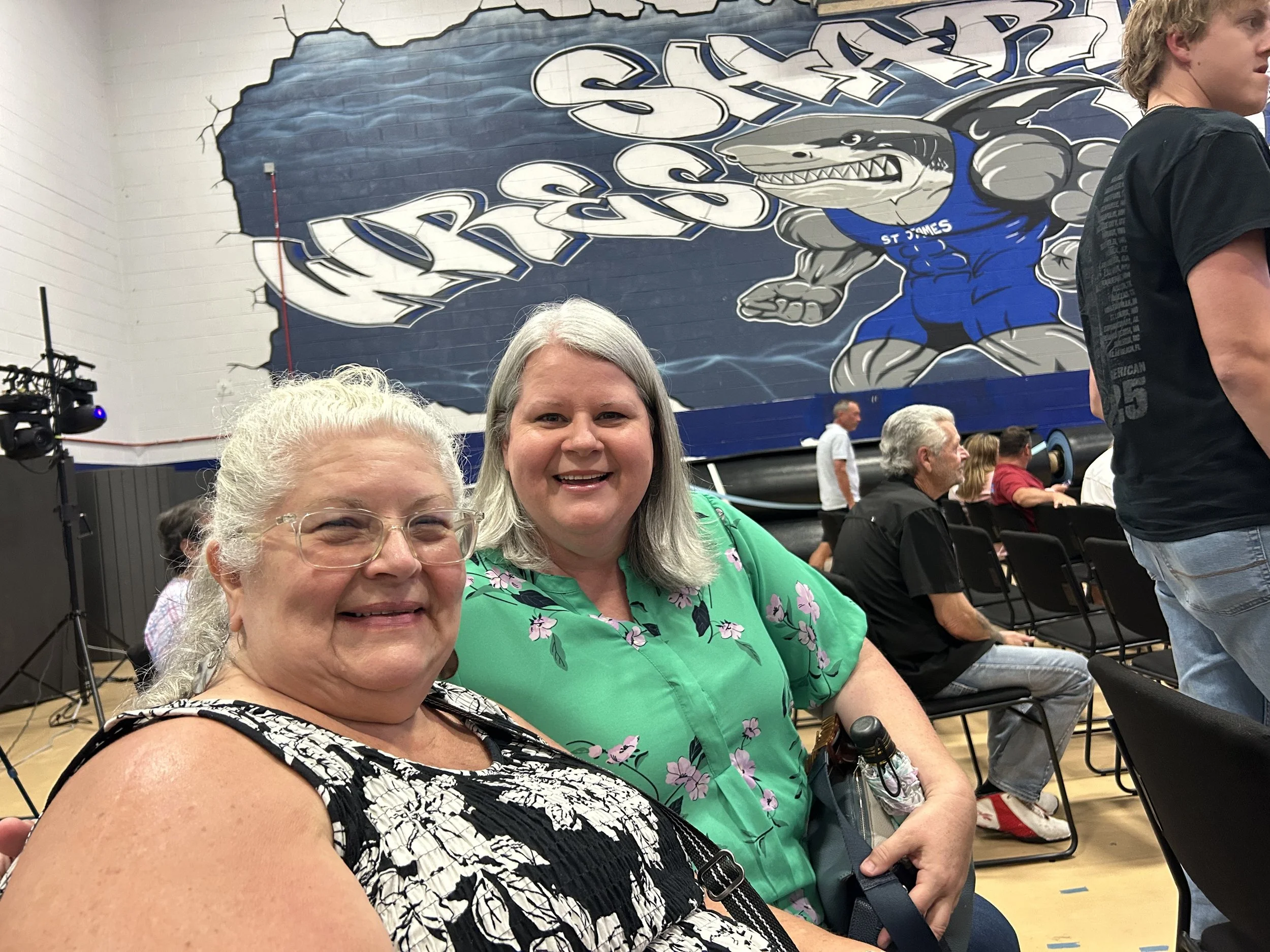 Two women smiling at the camera seated indoors during an event, with a large mural of a stylized alligator or crocodile and the words 'ST. JAMES' on the wall behind them, and other people seated in the background.