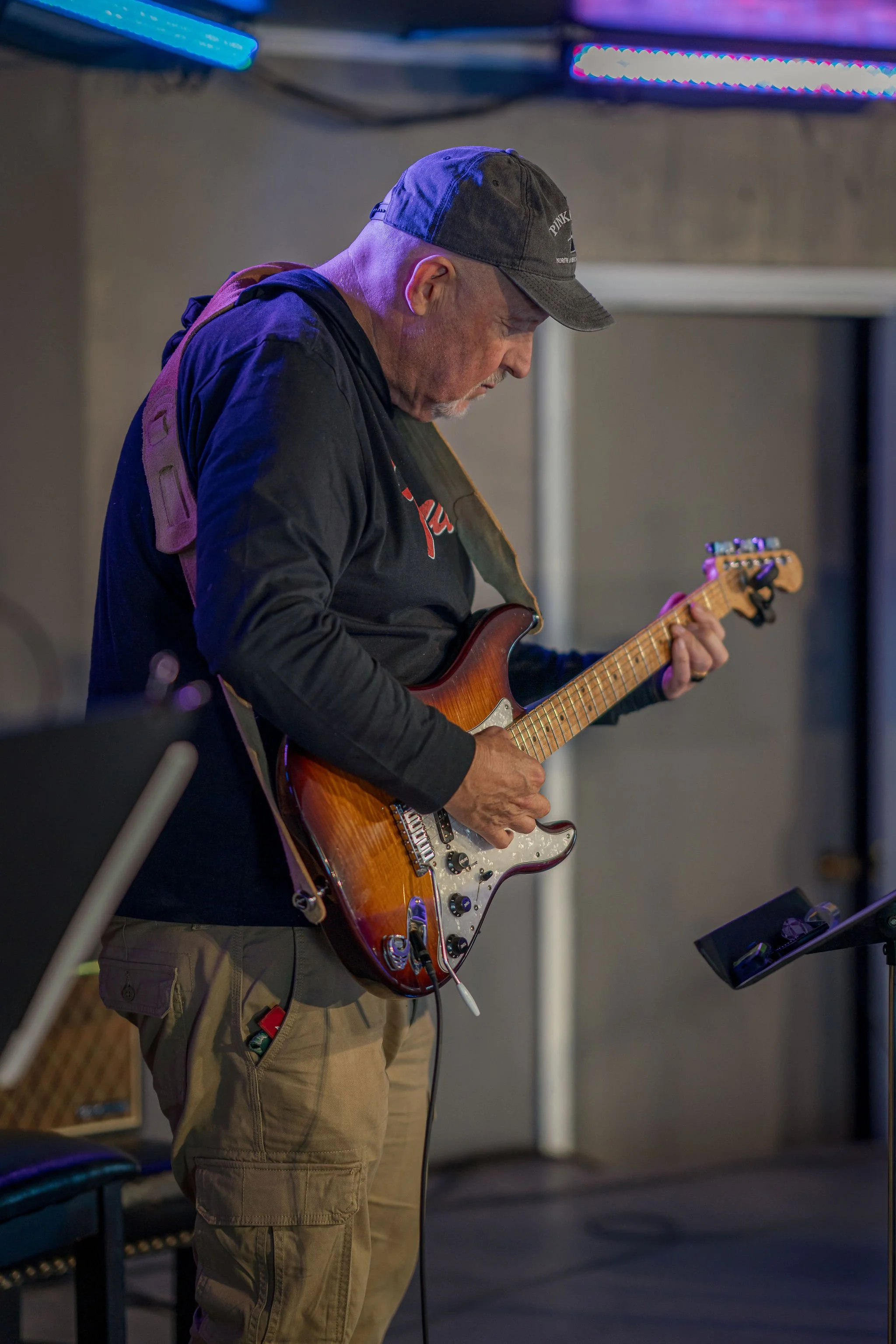 A man wearing a baseball cap and black jacket playing an electric guitar in a studio with gray walls and colorful LED lighting.