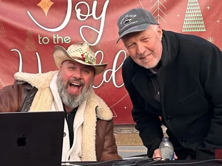 Two men smiling at the camera, one wearing a cowboy hat decorated with holly and a shear-lined jacket, the other in a dark jacket and cap, with a Christmas-themed backdrop that says "Joy to the World."